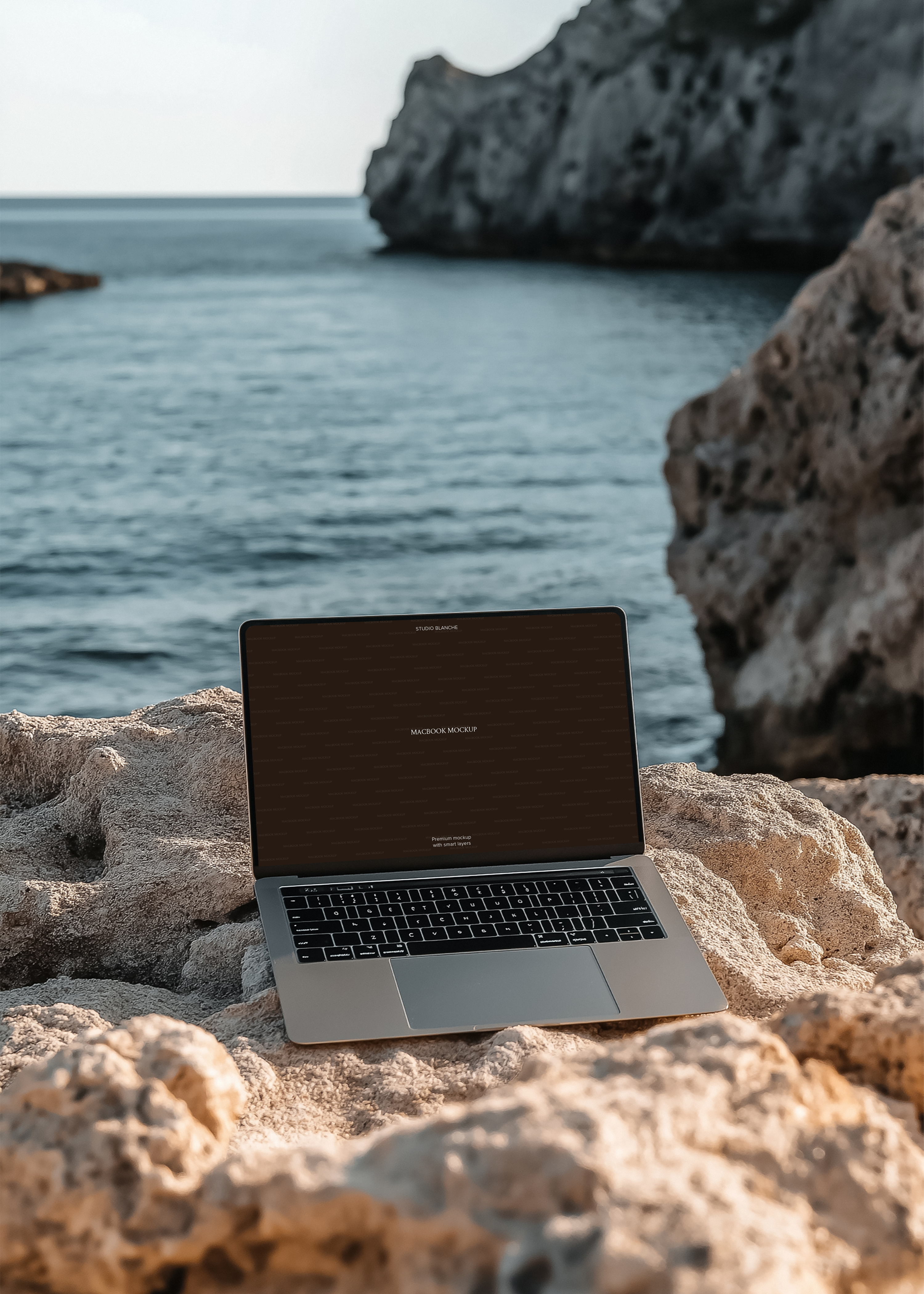 MacBook mockup with dark placeholder screen on rocky cove surface, still blue water and rock formations soft behind
