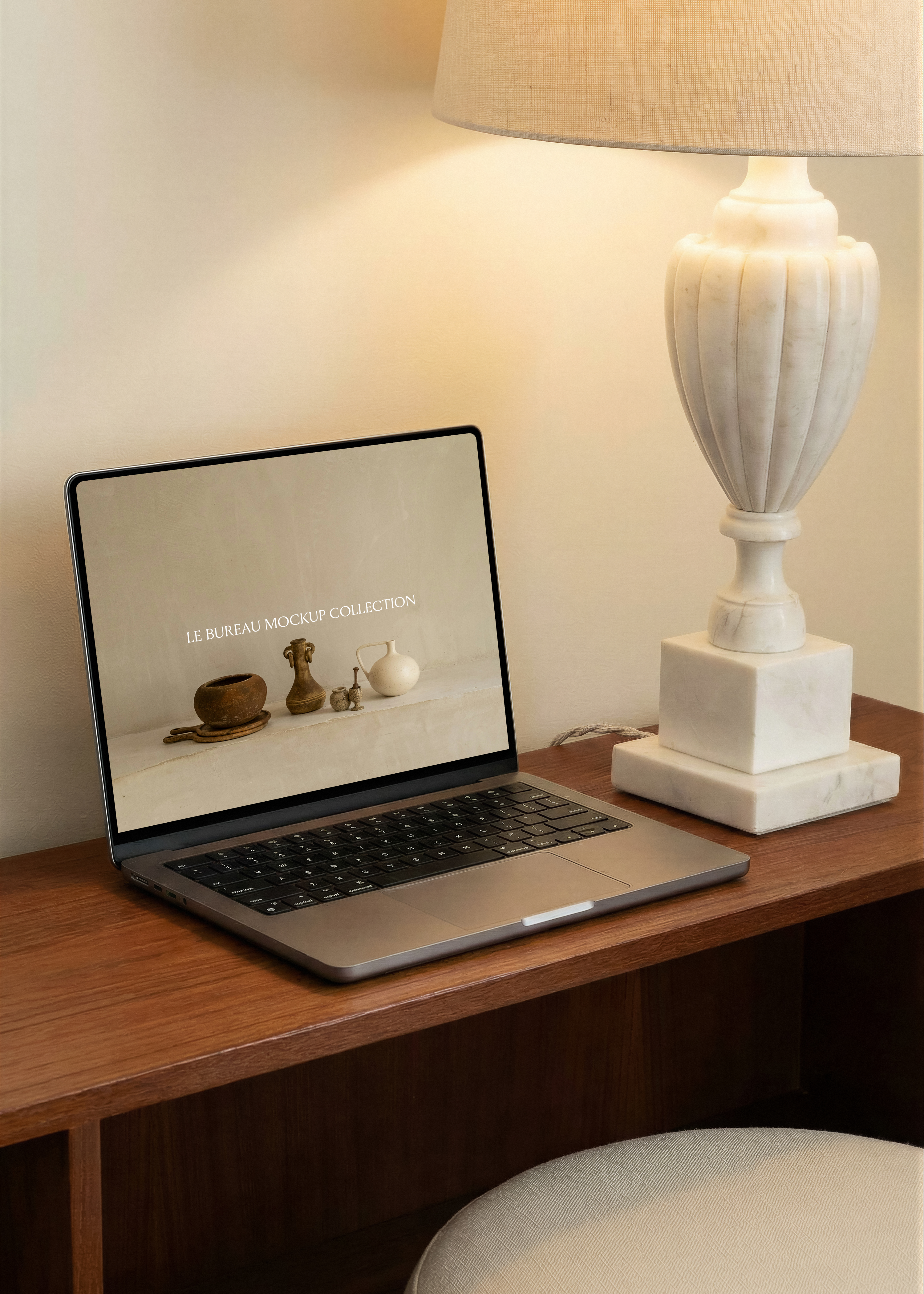 MacBook mockup displaying Le Bureau editorial on walnut console, carved marble urn lamp and linen shade warm interior