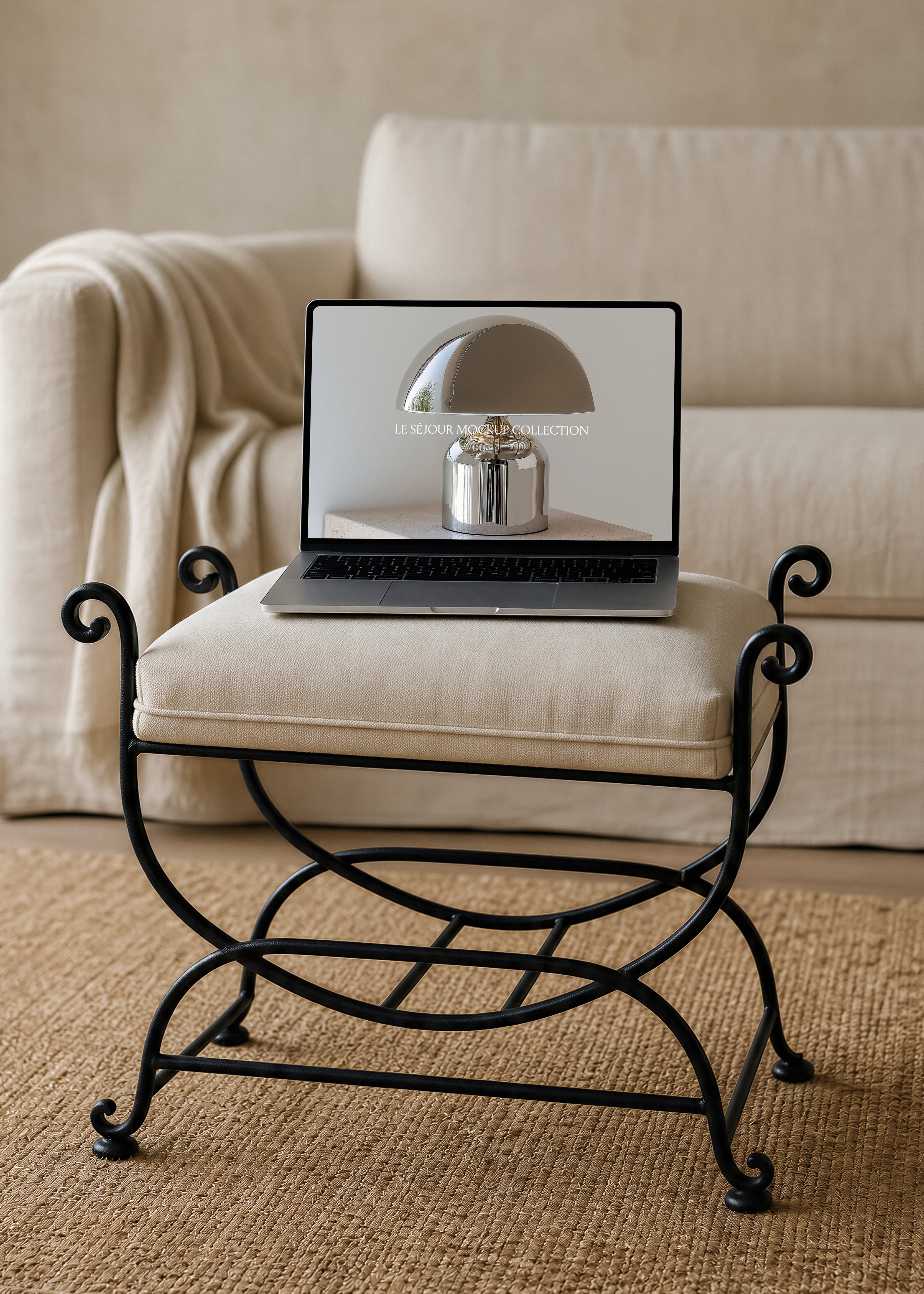 Open MacBook mockup on a linen cushioned wrought iron stool in front of a cream linen sofa with throw, woven jute rug below