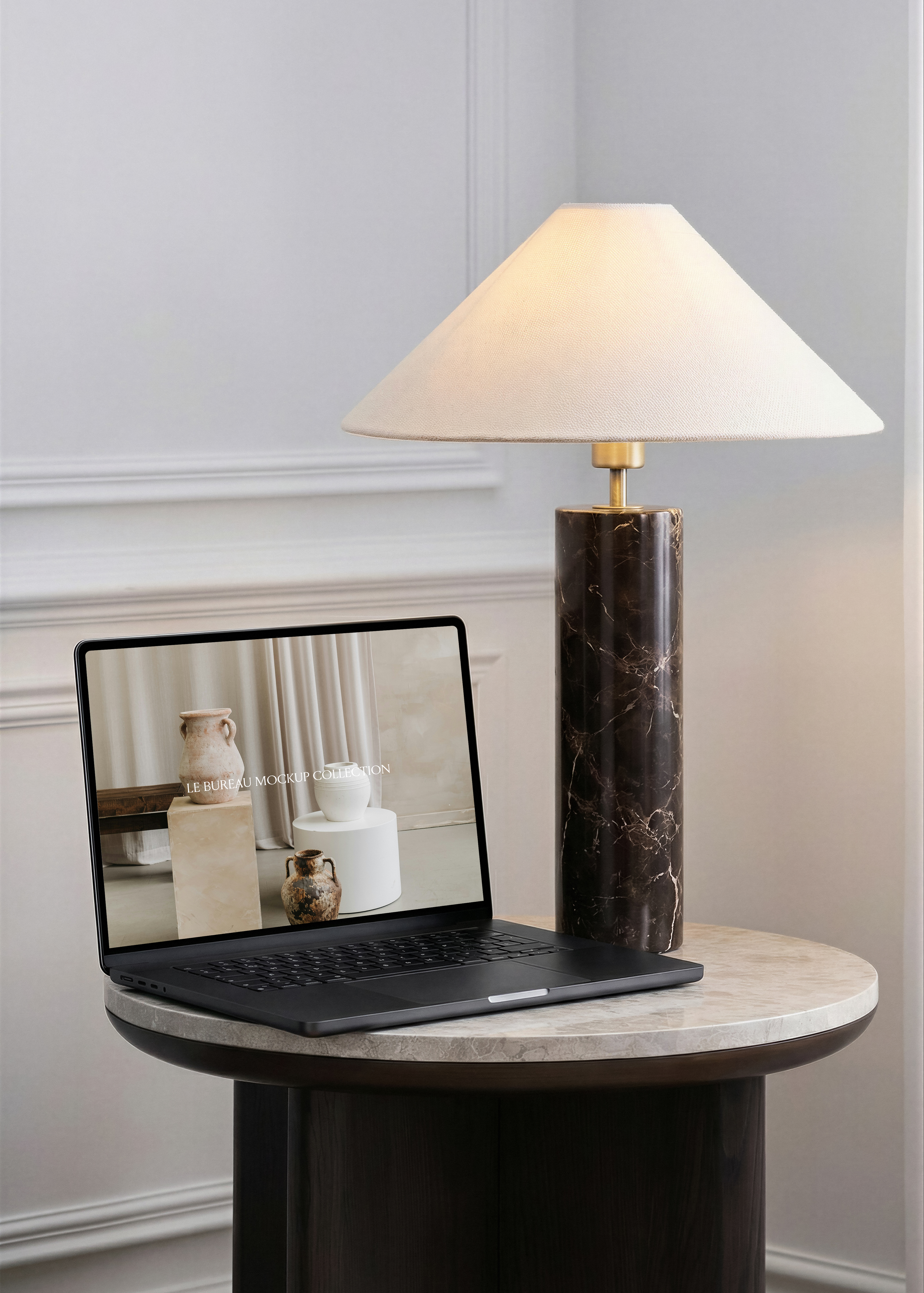 MacBook mockup displaying Le Bureau editorial on marble side table, dark marble column lamp and dove-grey panelled wall backdrop
