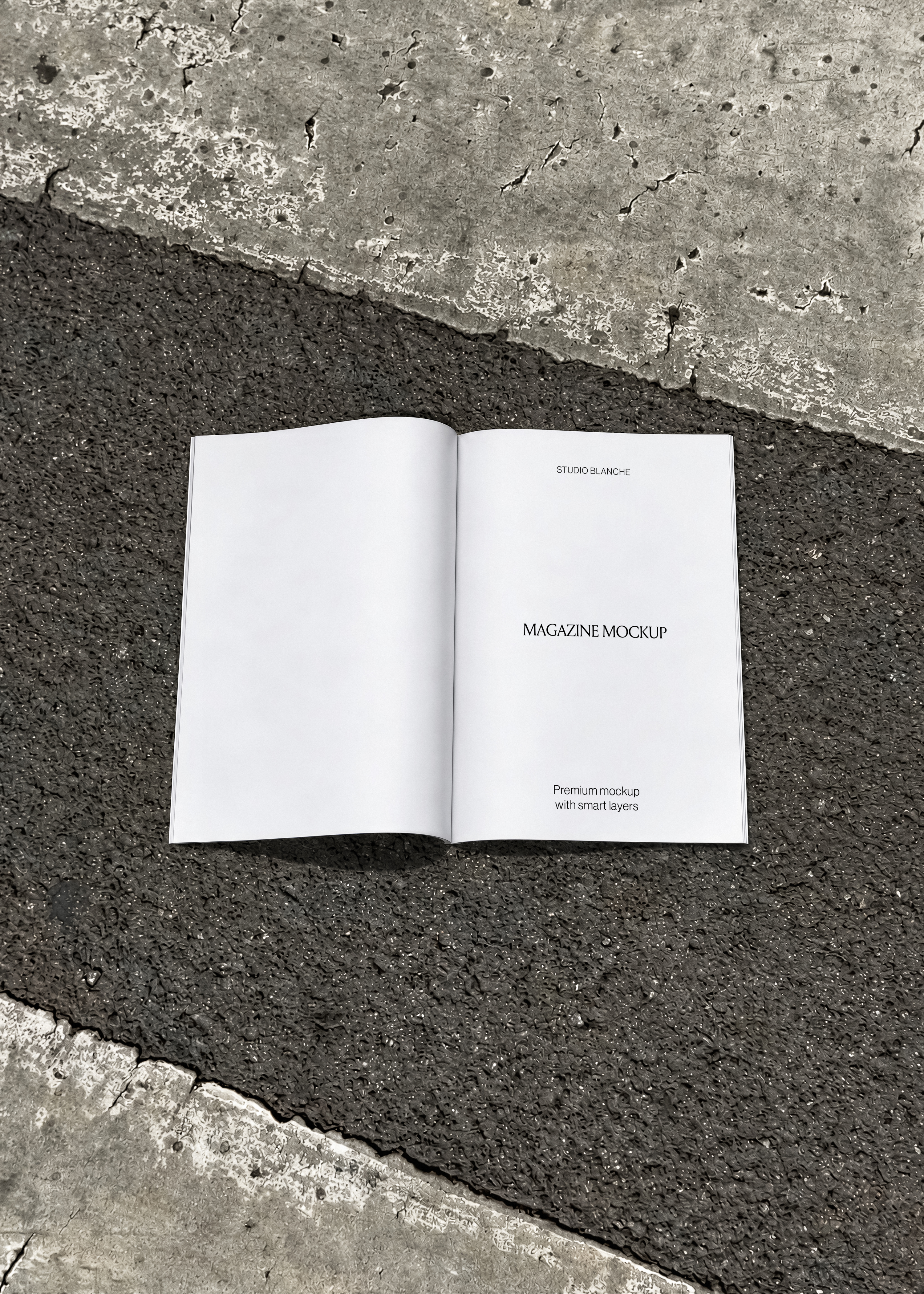Blank white magazine mockup open spread lying flat on dark asphalt strip between cracked grey concrete, overhead view