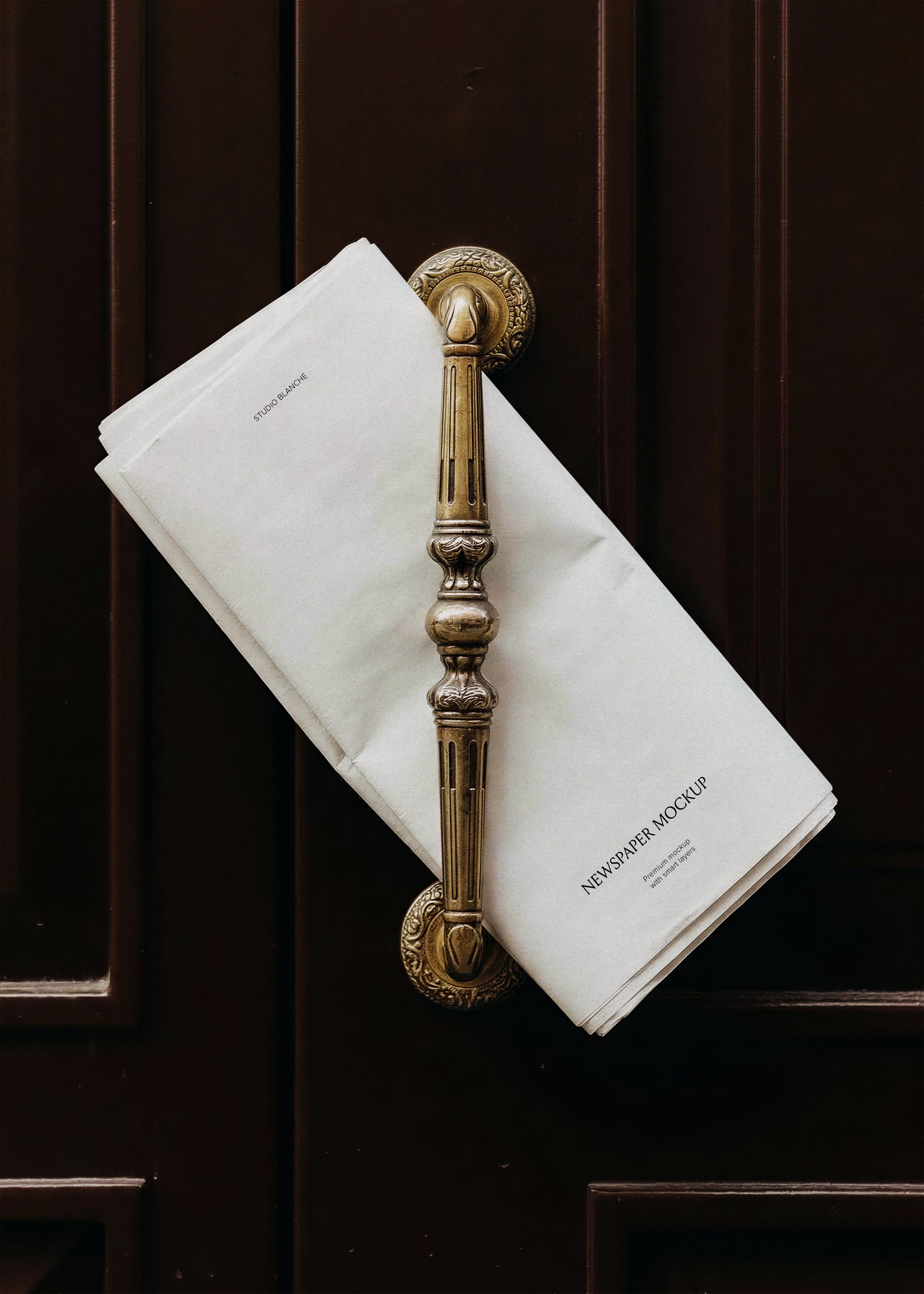 Blank folded newspaper mockup hanging on an ornate antique brass door handle against dark mahogany wood panelling, from Studio Blanche