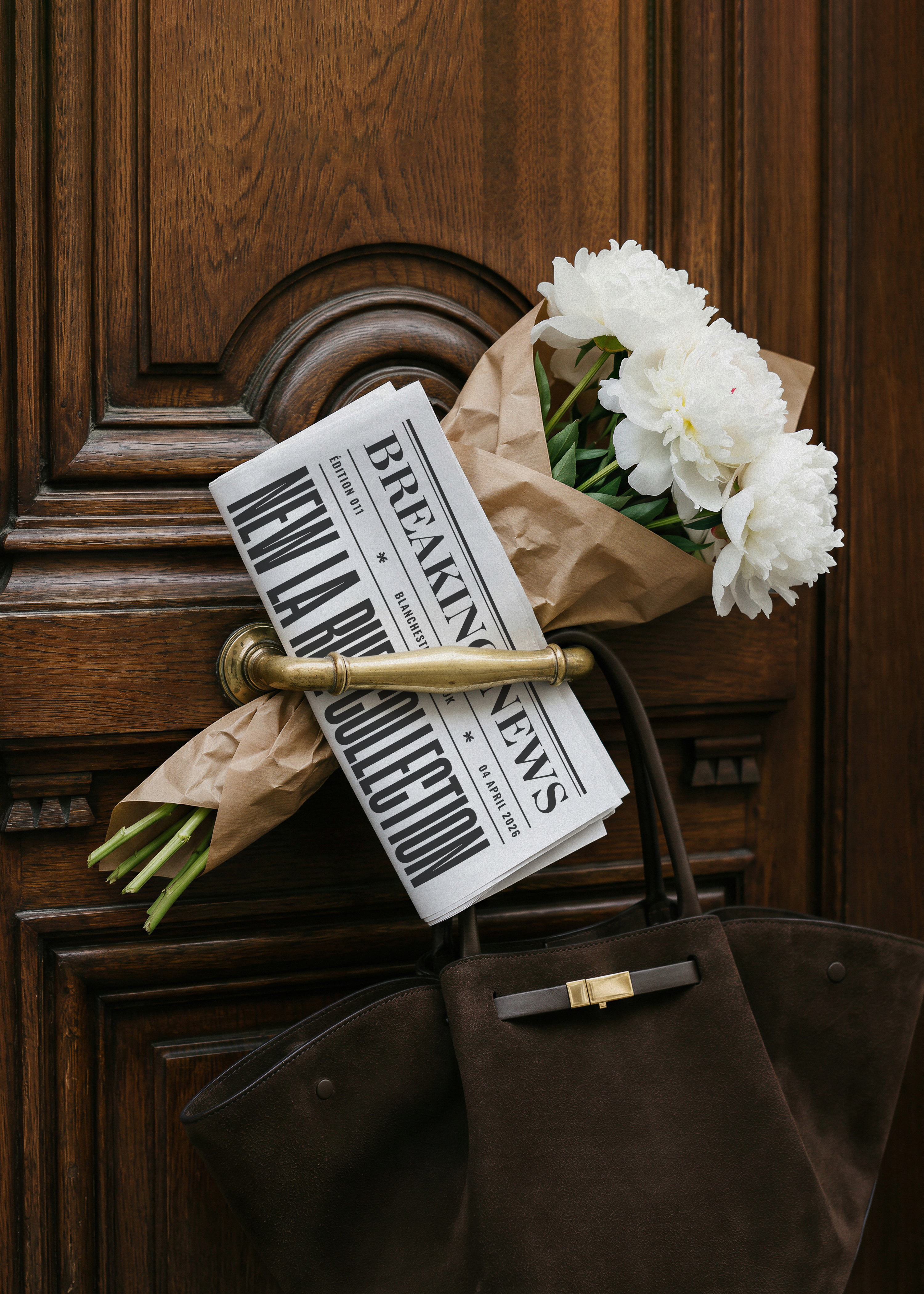 White newspaper mockup with minimal serif text tucked into ornate carved wooden door handle with bouquet and handbag