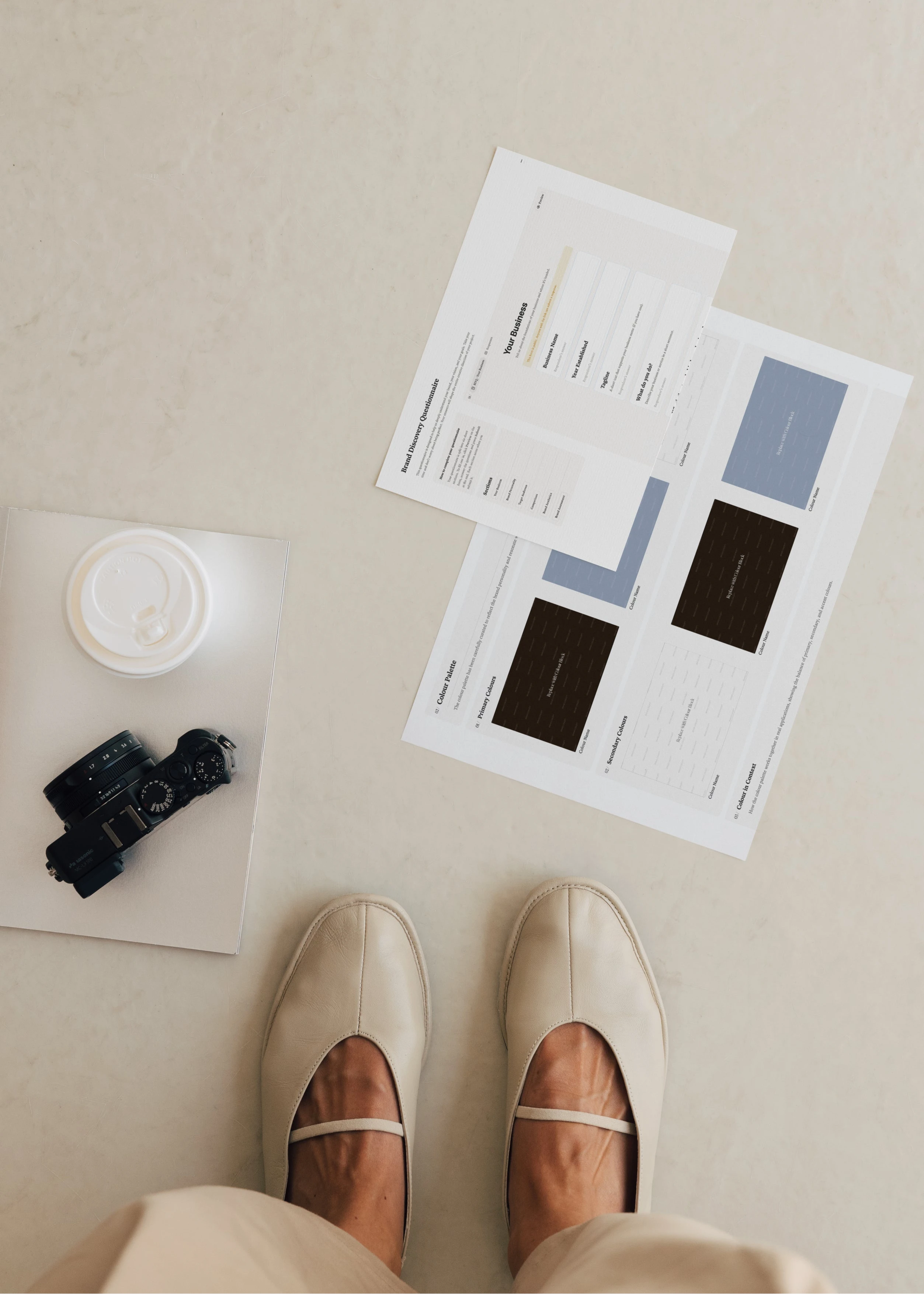 Printed pages from a Notion brand identity client portal showing the Brand Discovery Questionnaire and colour palette, on a stone floor with a camera and coffee.