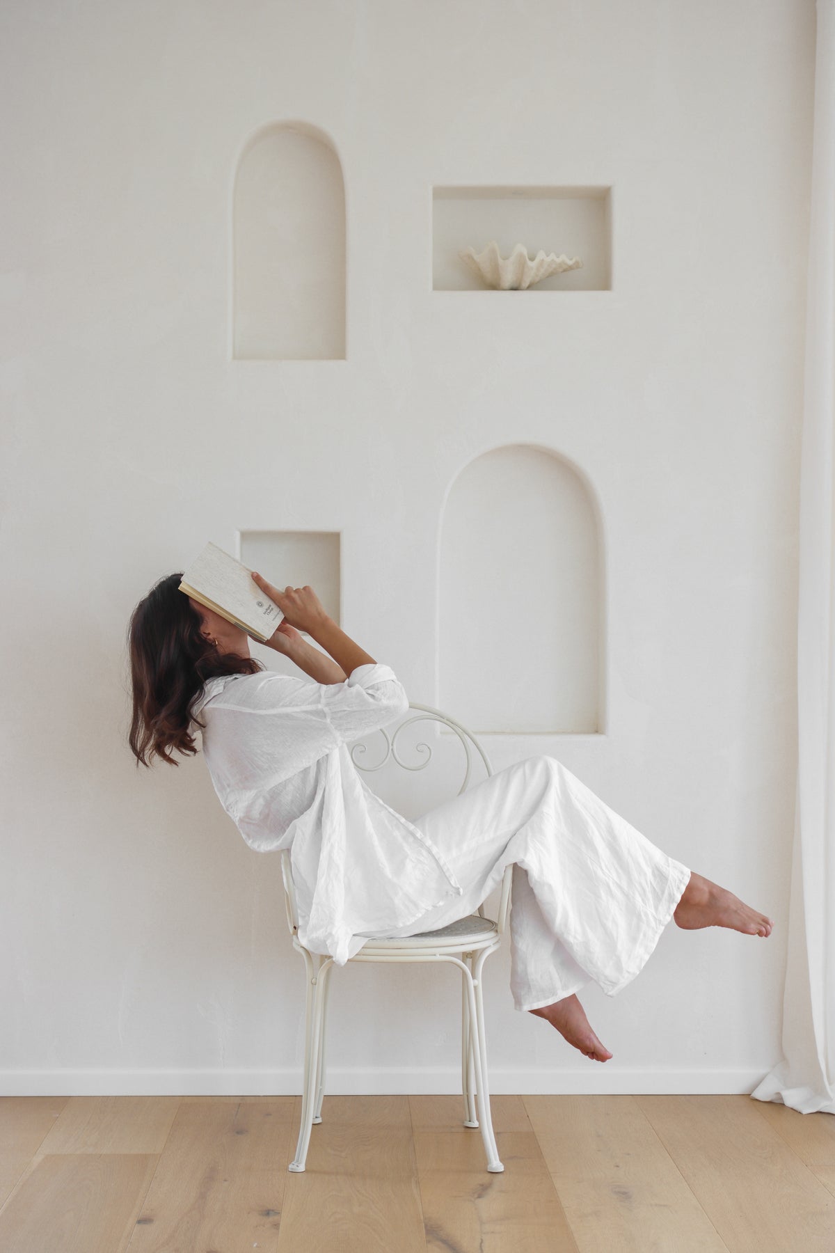 A woman in a white robe sits on a white chair, holding a book to her face in a bright, minimalist room with white walls