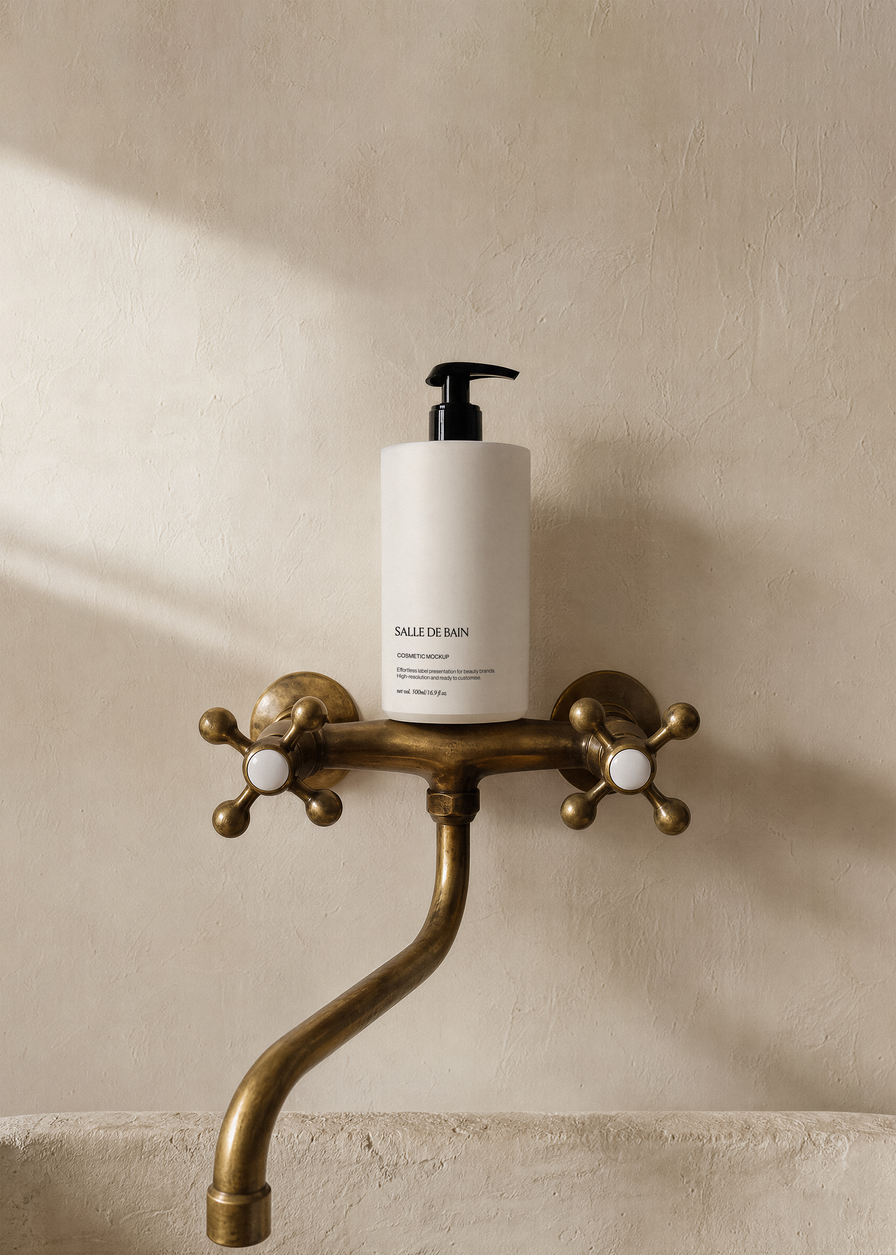 White cosmetic pump bottle mockup with black pump balanced on antique brass cross tap, warm plaster wall and soft daylight
