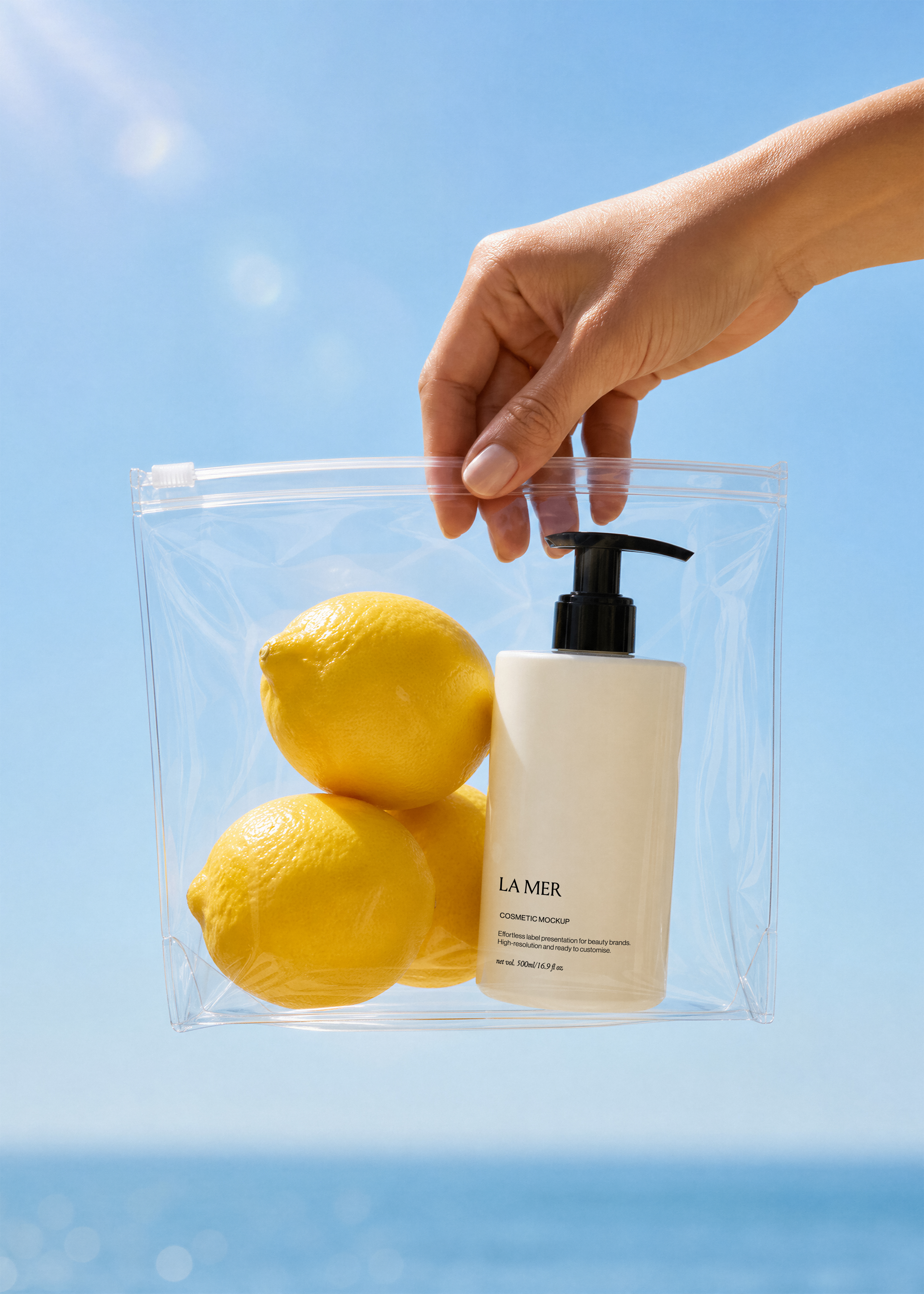 White cosmetic pump bottle mockup and three lemons inside a clear zip bag held by a hand against a blue coastal sky