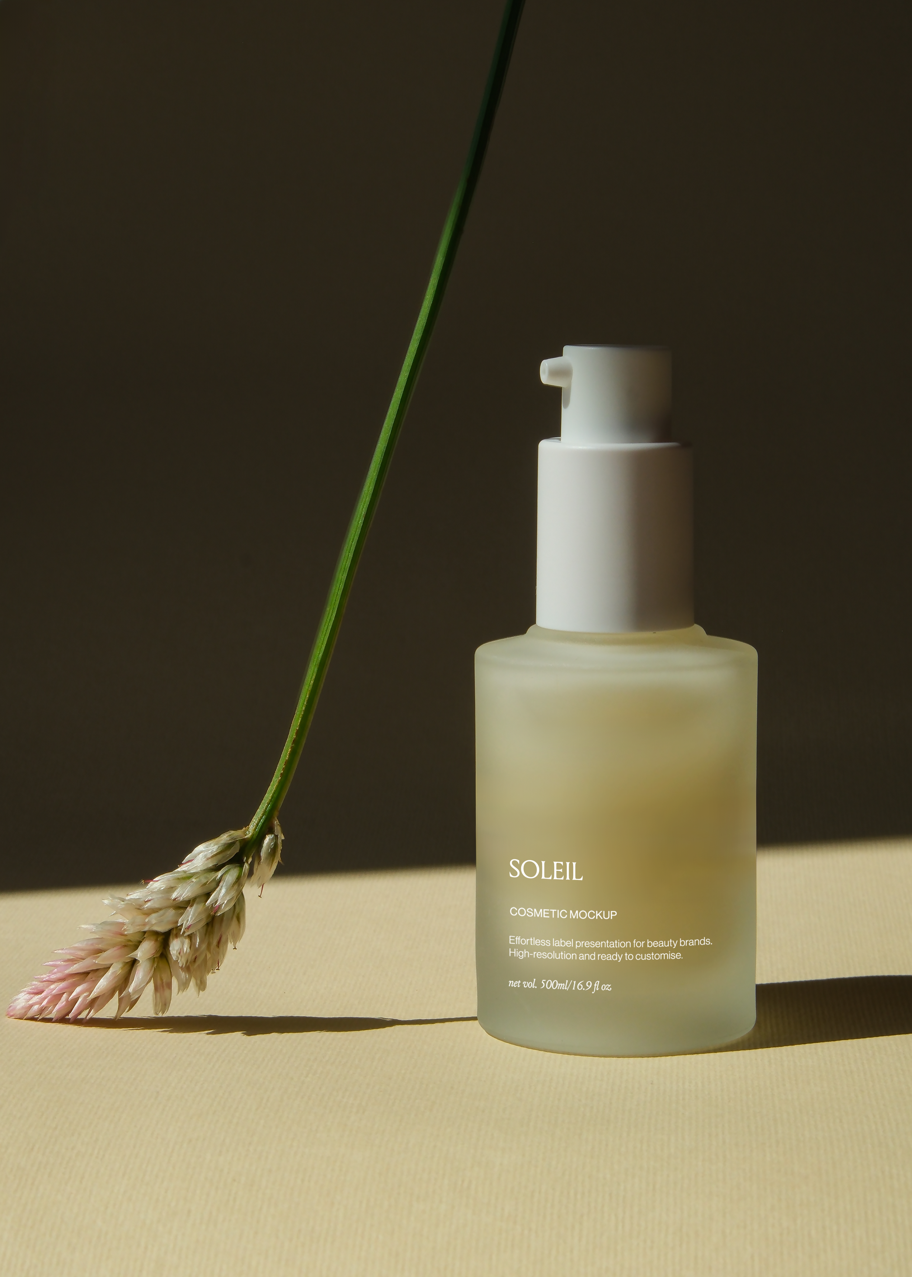 Frosted glass pump bottle mockup on sand-toned surface beside dried grass stem, dark backdrop, warm directional light