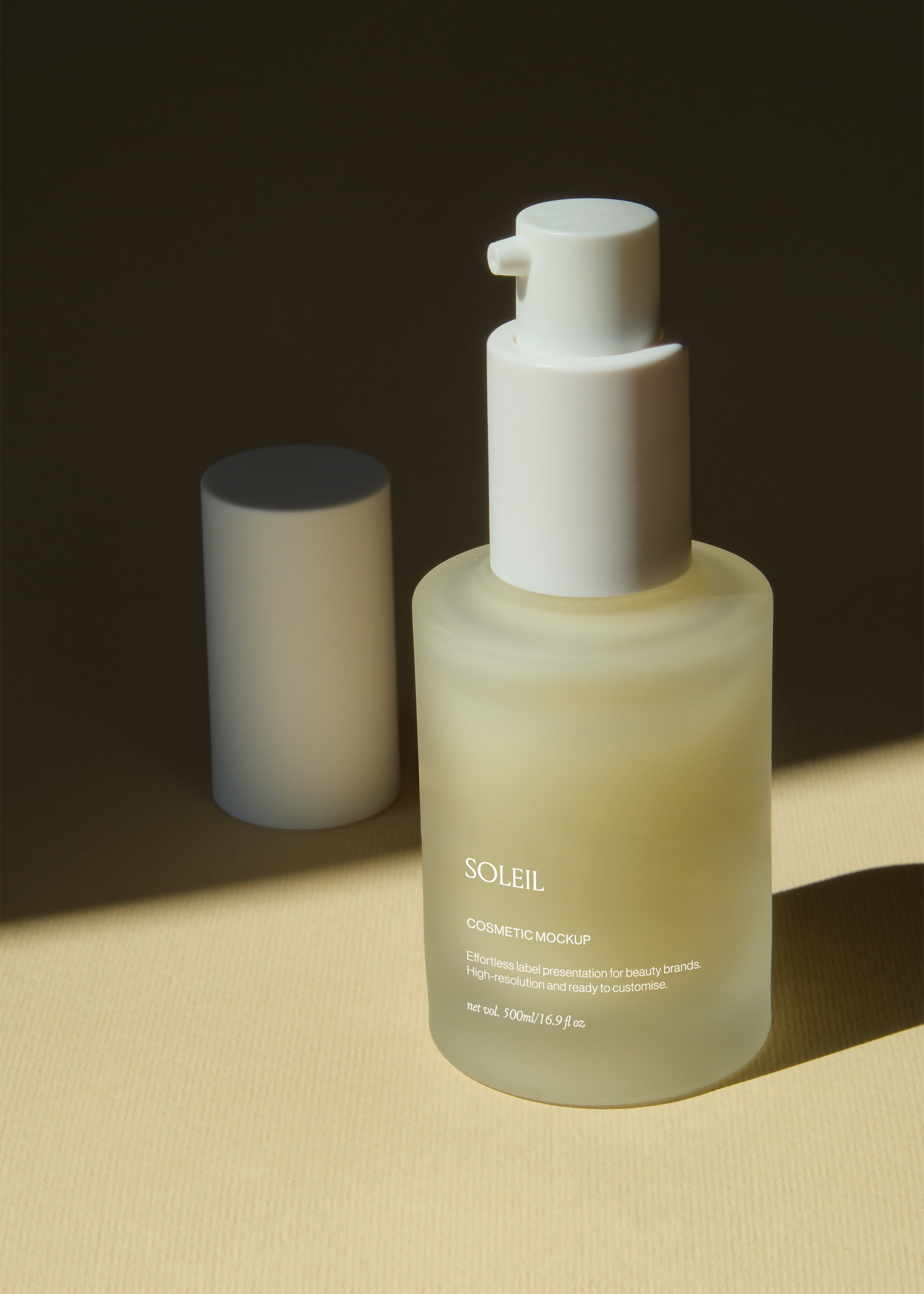 Frosted glass pump bottle mockup beside cylindrical prop on sand-toned surface, dark backdrop, warm directional light