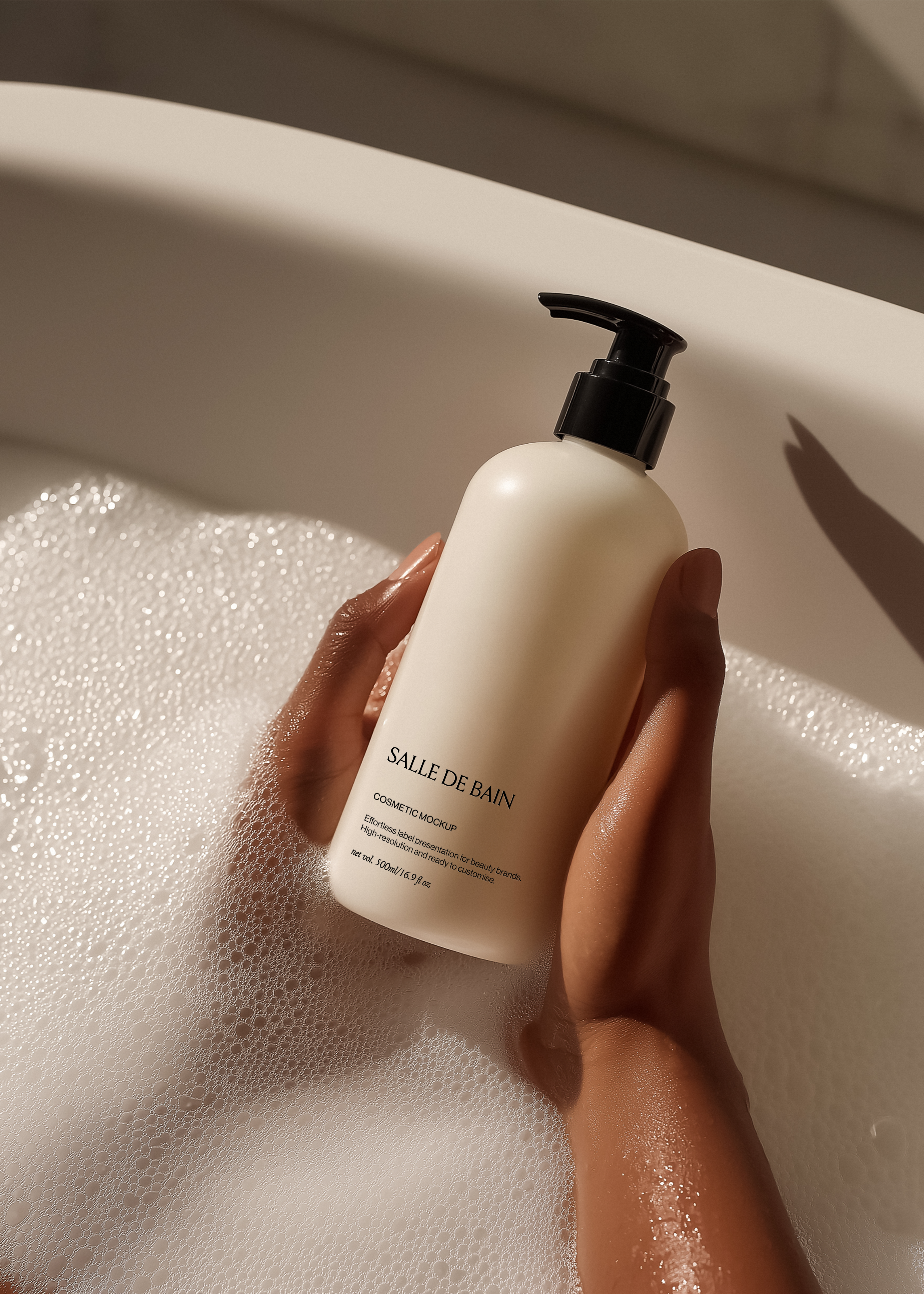 Hand holding a cream cosmetic pump bottle with black dispenser above a foamy bubble bath in warm directional light