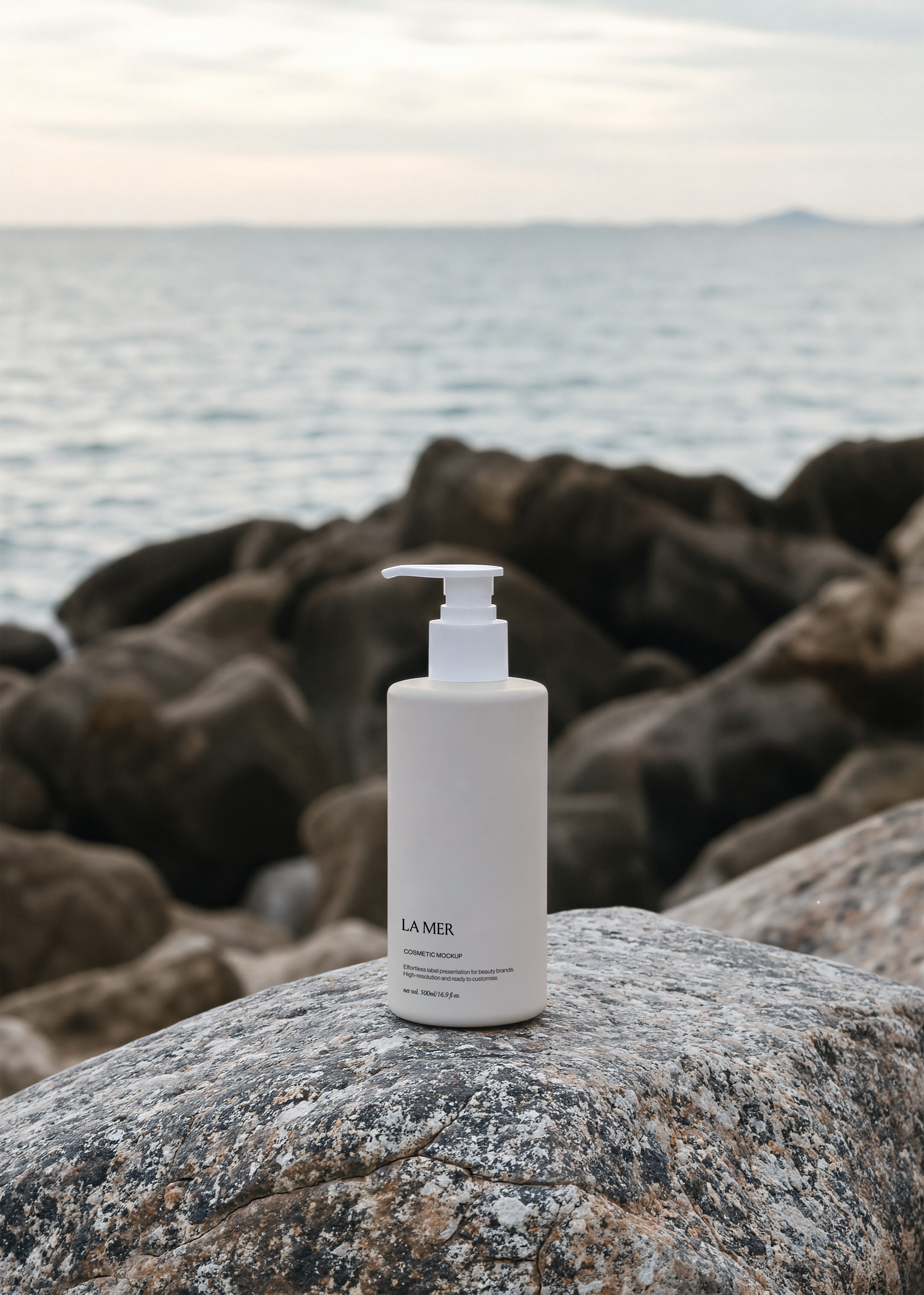 Tall white cosmetic pump bottle on smooth granite rock, dark coastal rocks and still silver sea blurred behind