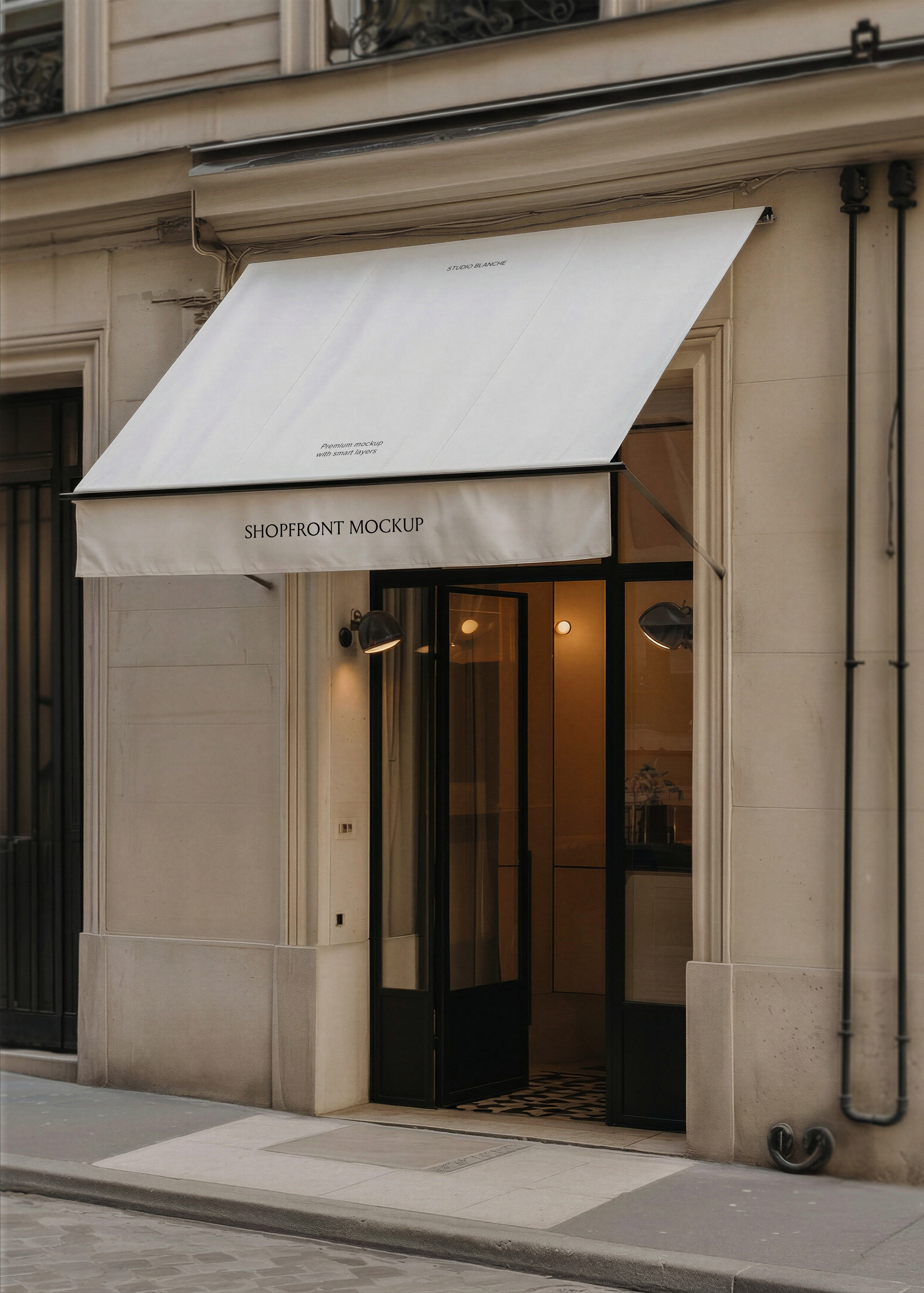 Blank white canvas shopfront awning mockup on a Parisian limestone building facade with open black-framed glass door and warm interior lighting, from Studio Blanche