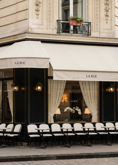 Shopfront mockup on Parisian café facade, cream canvas awning with branded lettering, black lacquered frontage, bistro chairs, stone building
