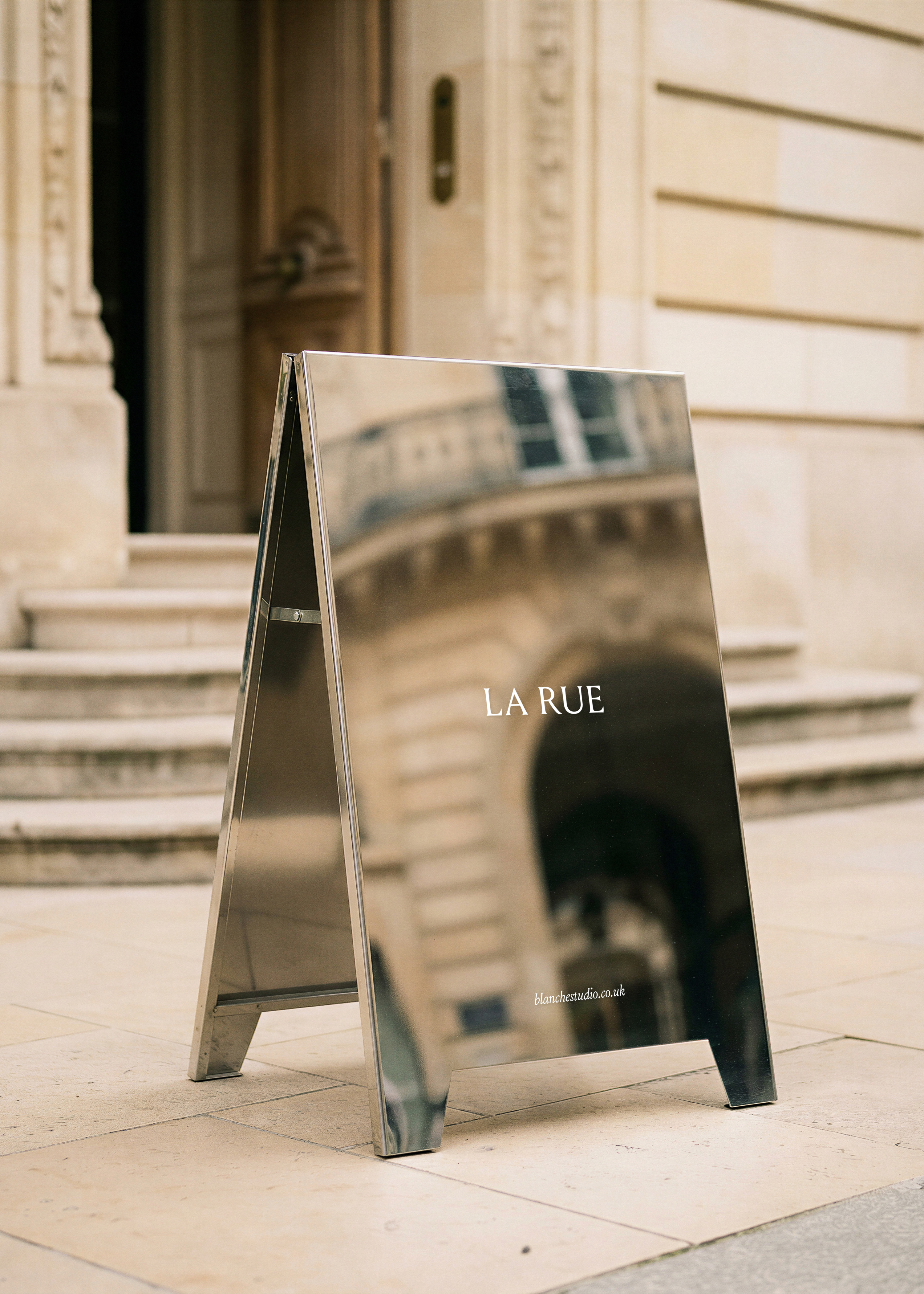 Polished steel sandwich board mockup with white serif text standing on limestone street outside ornate classical entrance