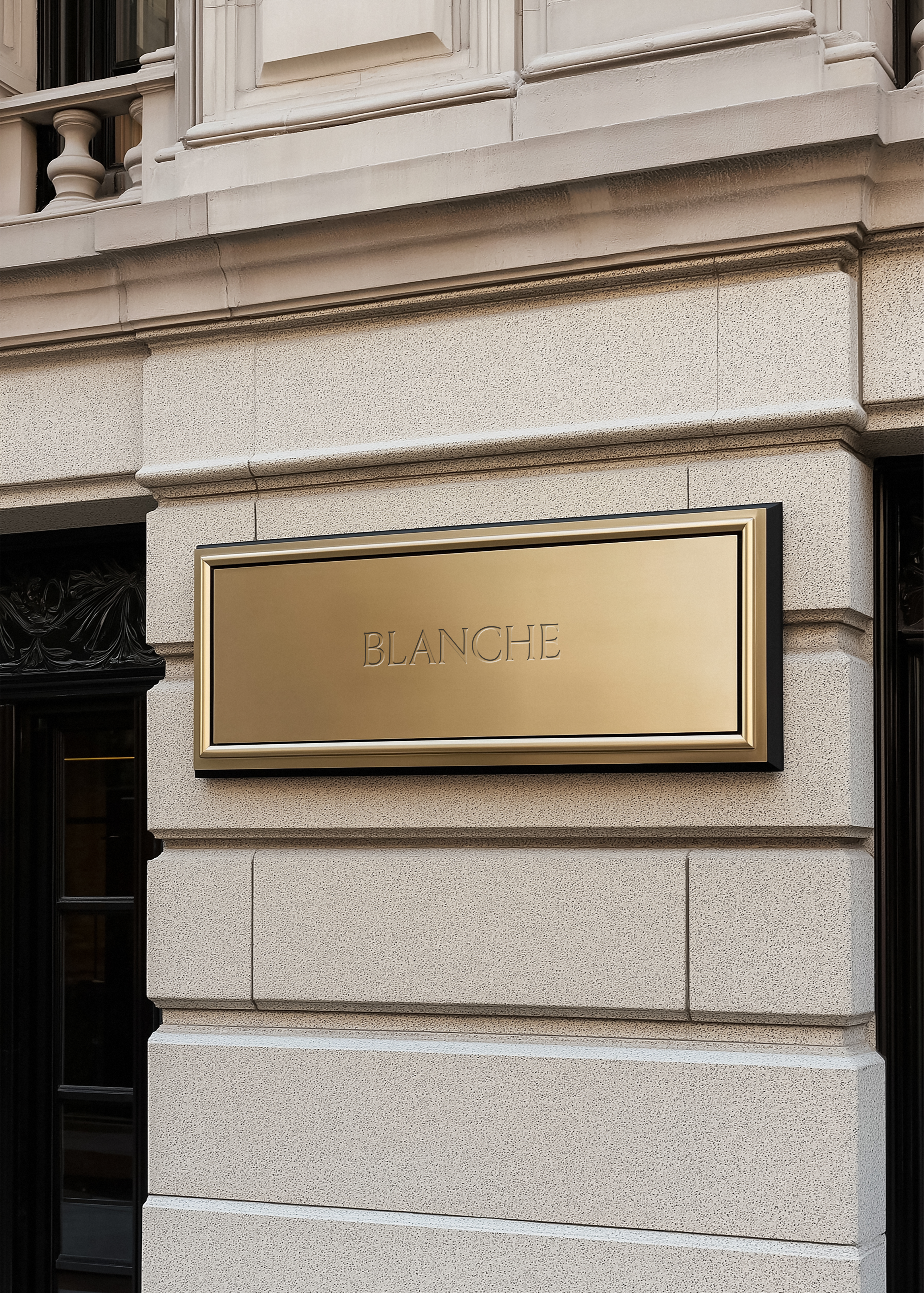 Gold metal sign mockup with engraved serif brand name in brass frame, pale limestone shopfront and dark doorway beside