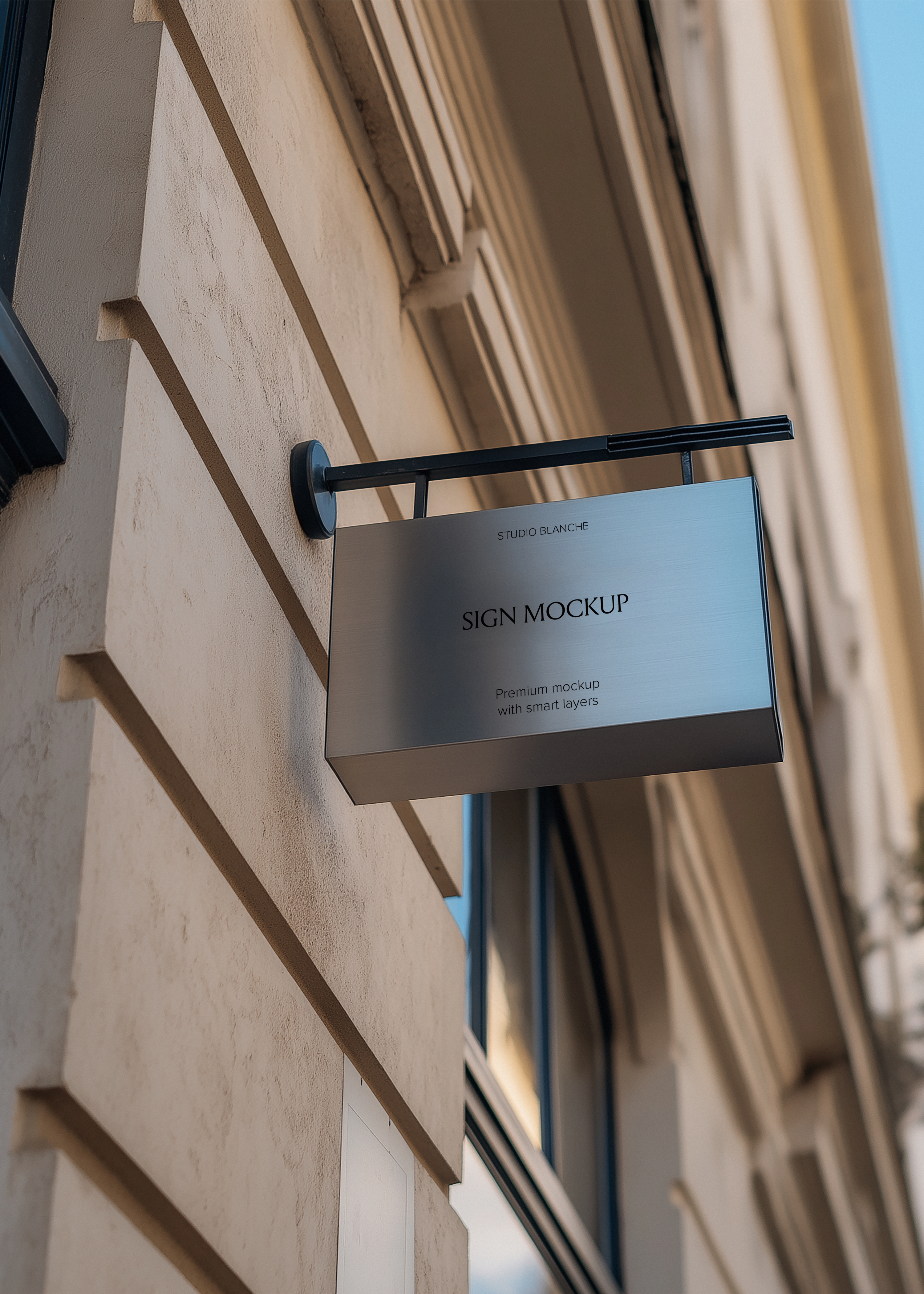 Brushed steel hanging sign mockup on black iron wall bracket, warm stone building facade and architectural cornice above
