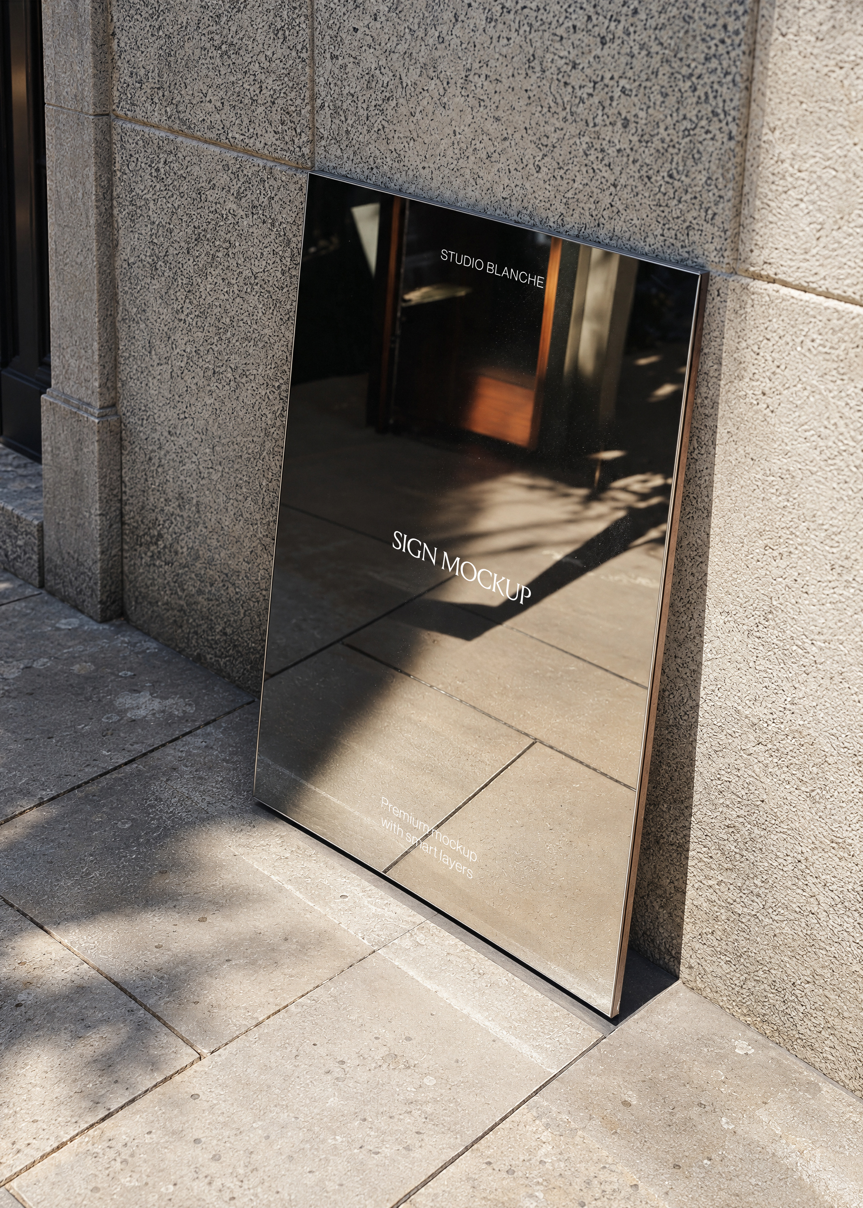 Dark mirror-finish sign mockup leaning against stone wall on tiled pavement, reflective surface, bronze trim detail