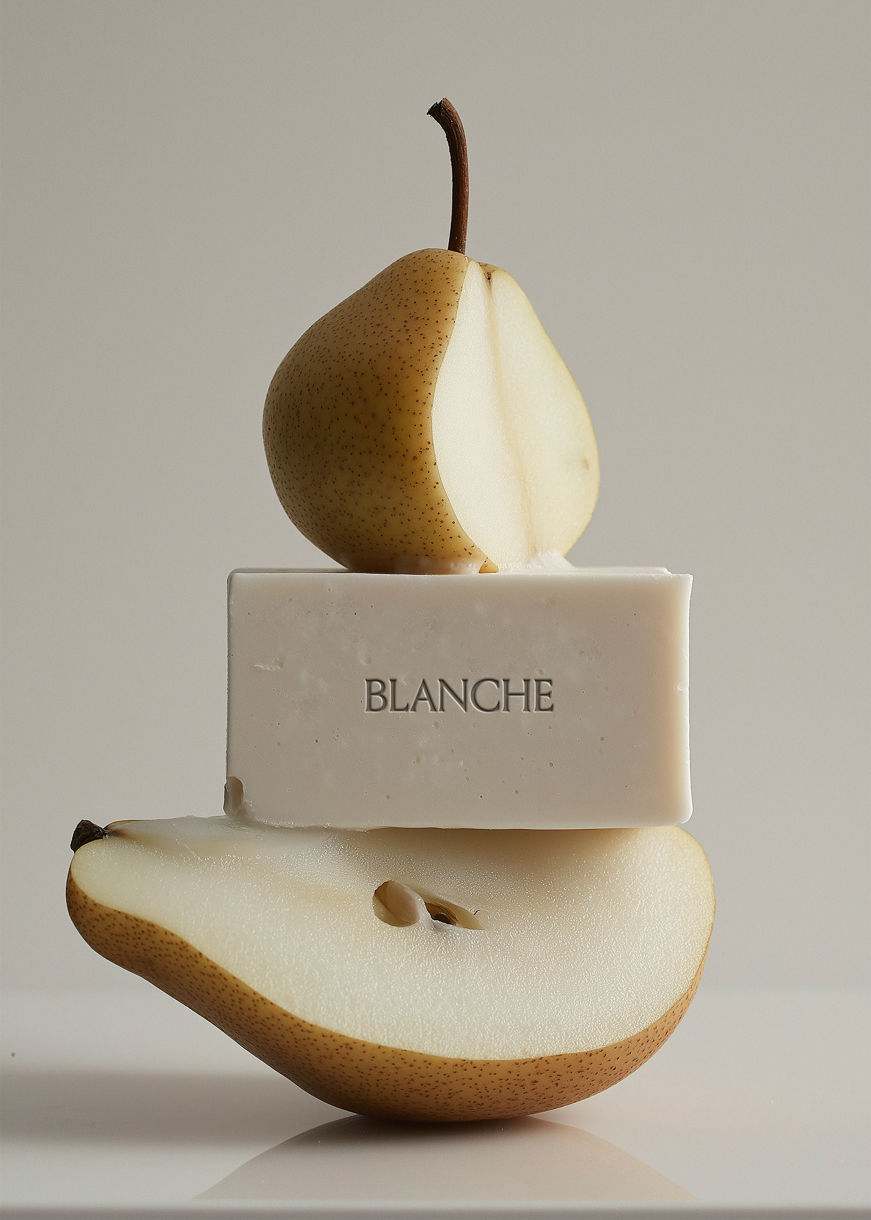 White soap bar sandwiched between two pear halves in stacked composition, grey background, embossed brand label, editorial styling
