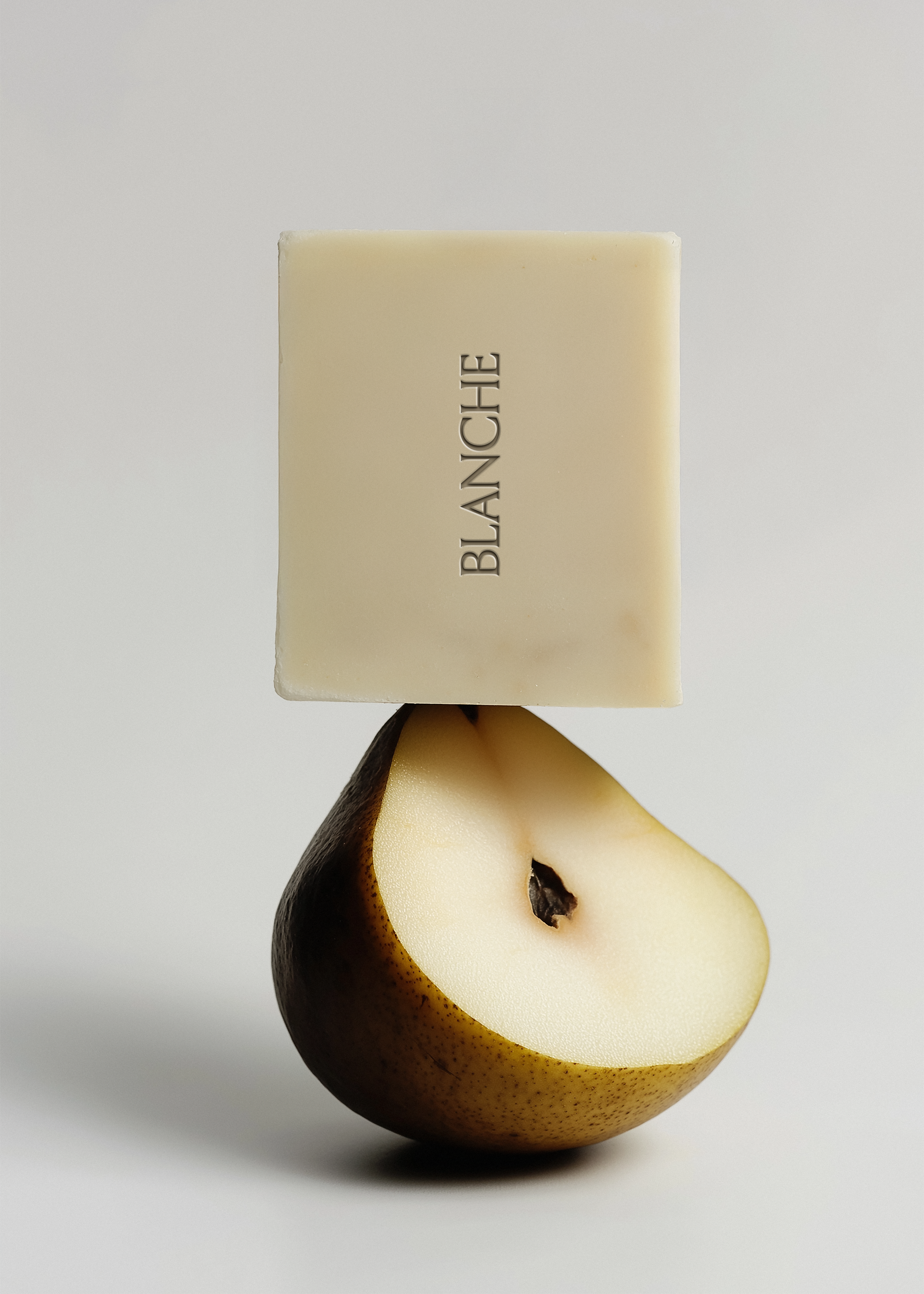 Smooth ivory square soap bar balanced on dark-skinned halved pear, light grey background, embossed vertical brand label