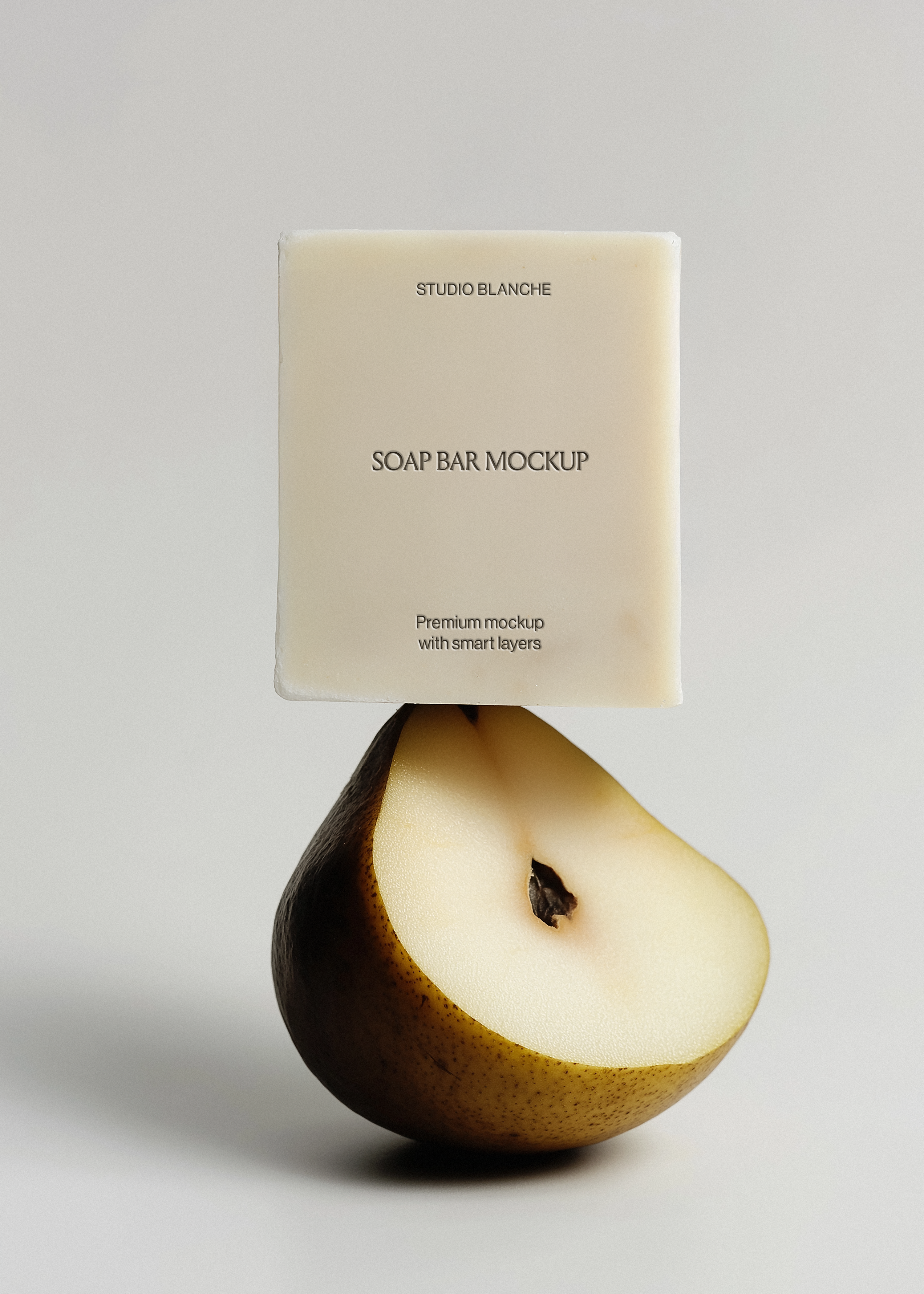 Clean ivory soap bar mockup on dark halved pear, Studio Blanche label placeholder, soft grey backdrop