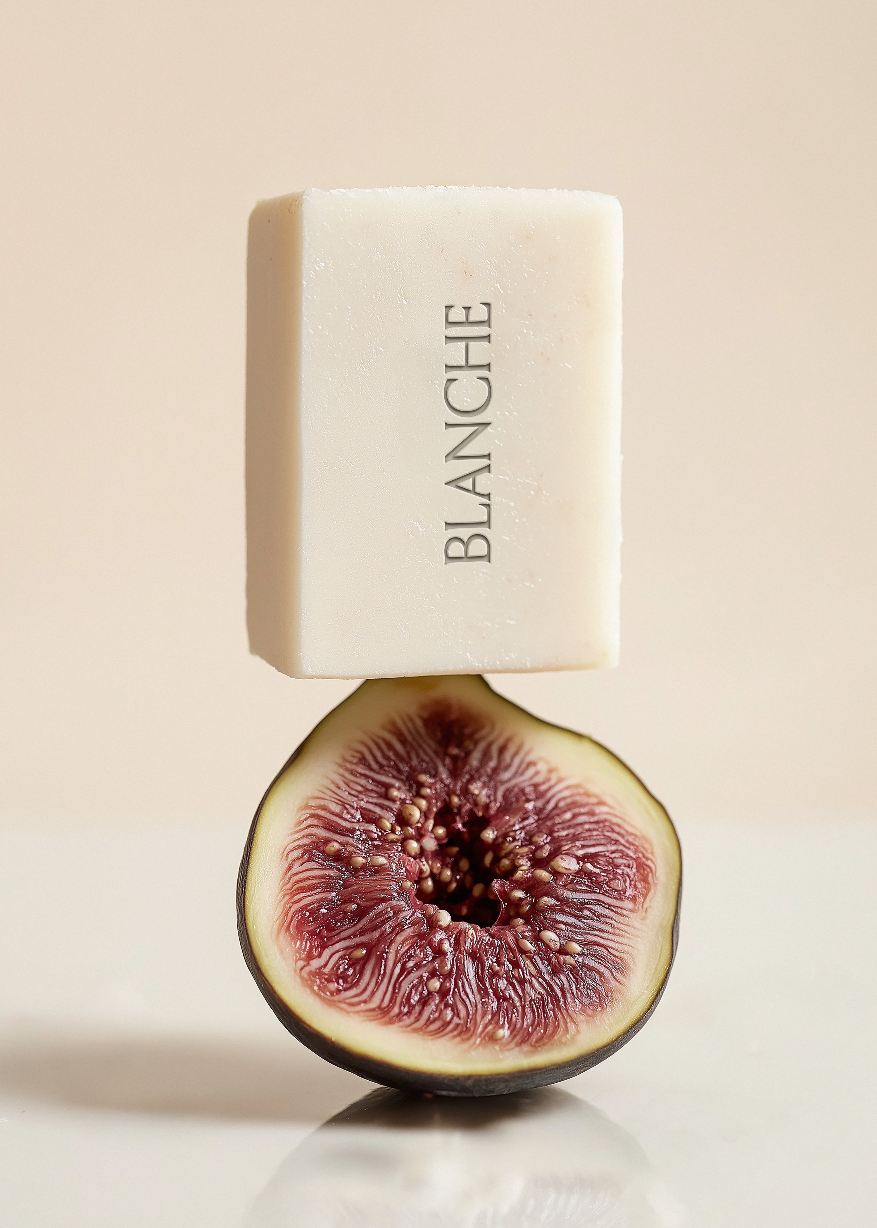 White textured square soap bar balanced on halved fig, blush background, deep crimson fruit interior, embossed brand label