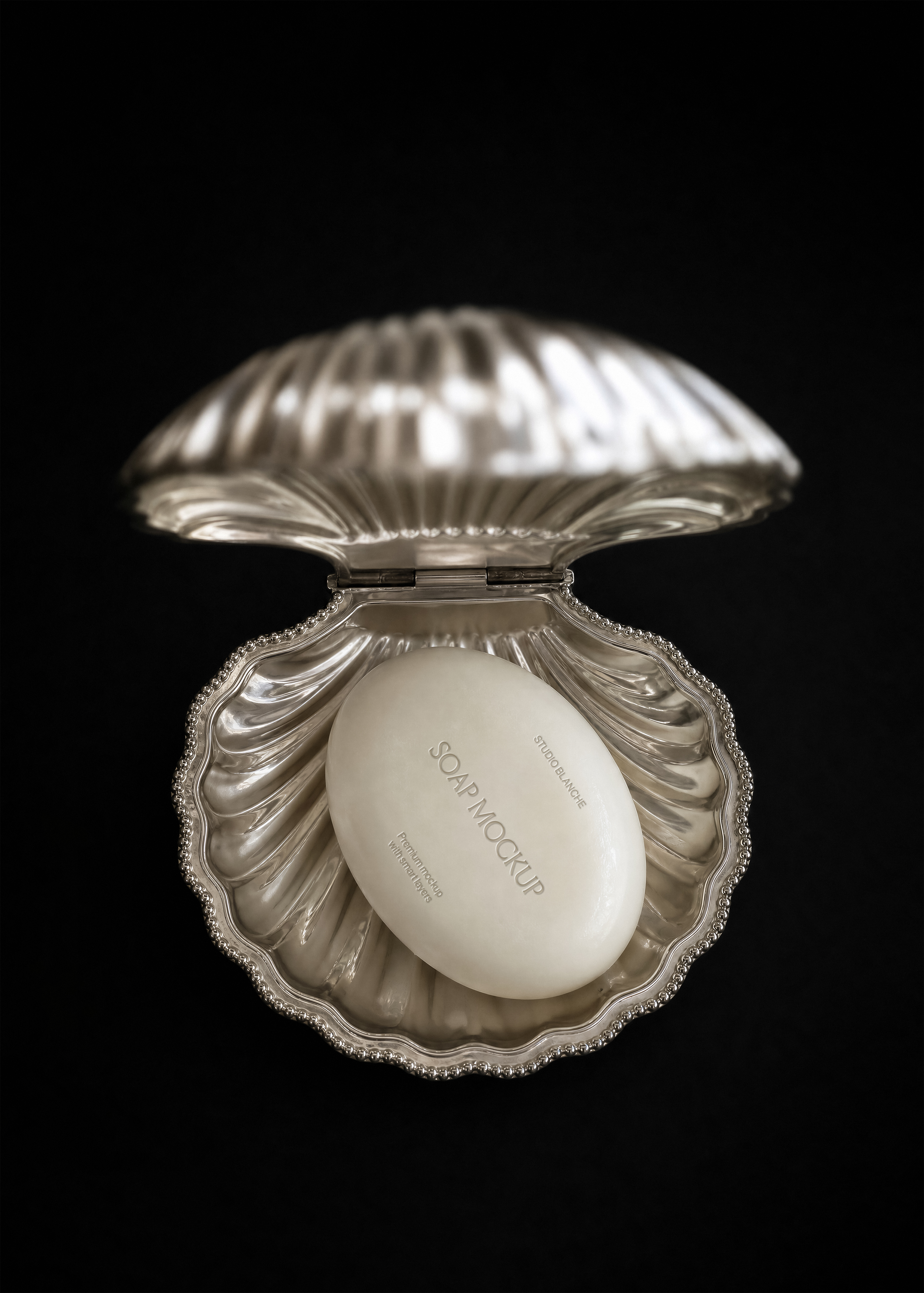 Detail view of an oval soap bar mockup in a hinged silver shell dish with beaded edge, dark studio backdrop