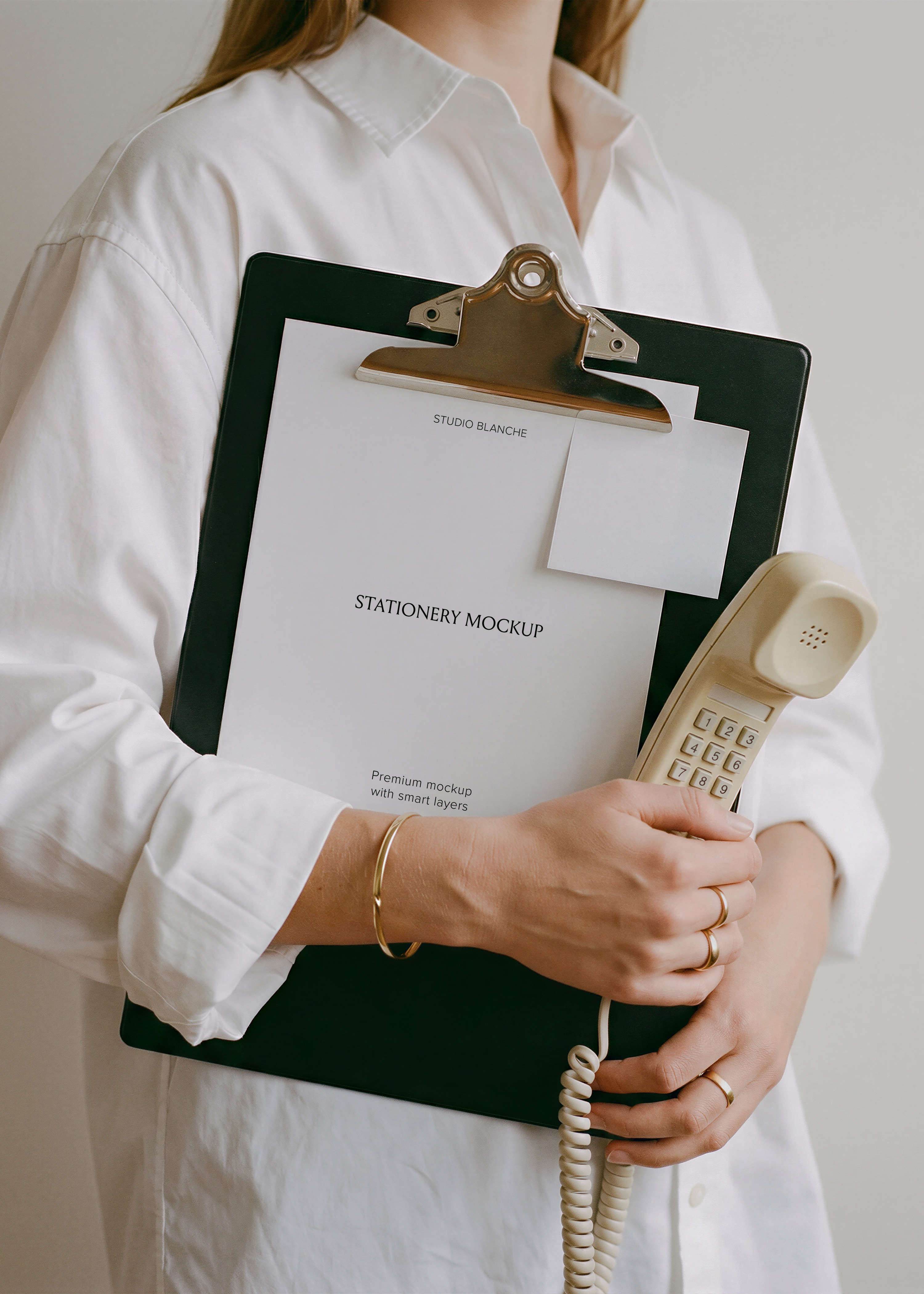 Blank letterhead with small paper note on black clipboard, cream corded phone handset tucked in arm, gold jewellery