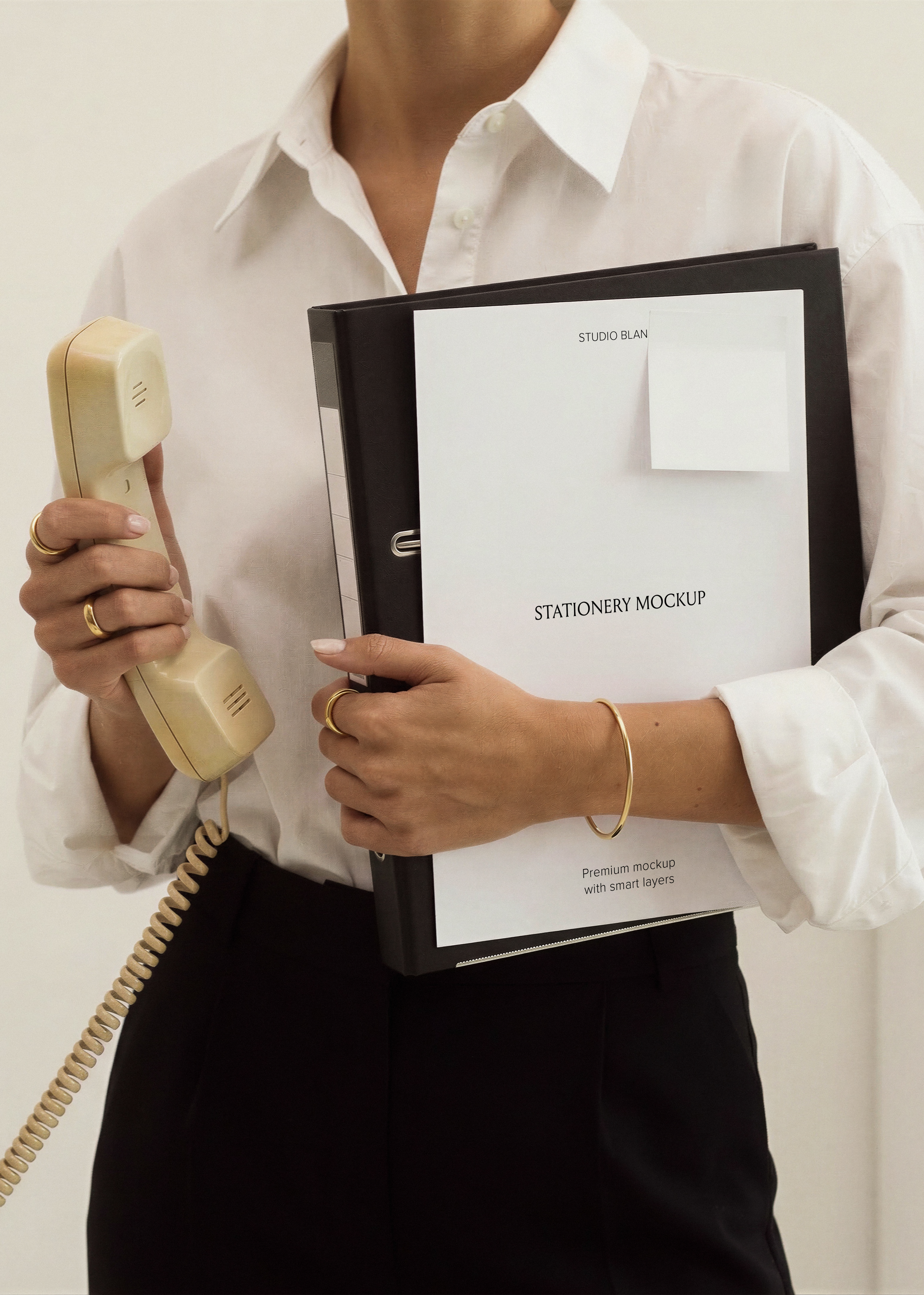 Blank A4 letterhead and small paper note on black leather binder, cream corded phone in hand, gold bangle and jewellery