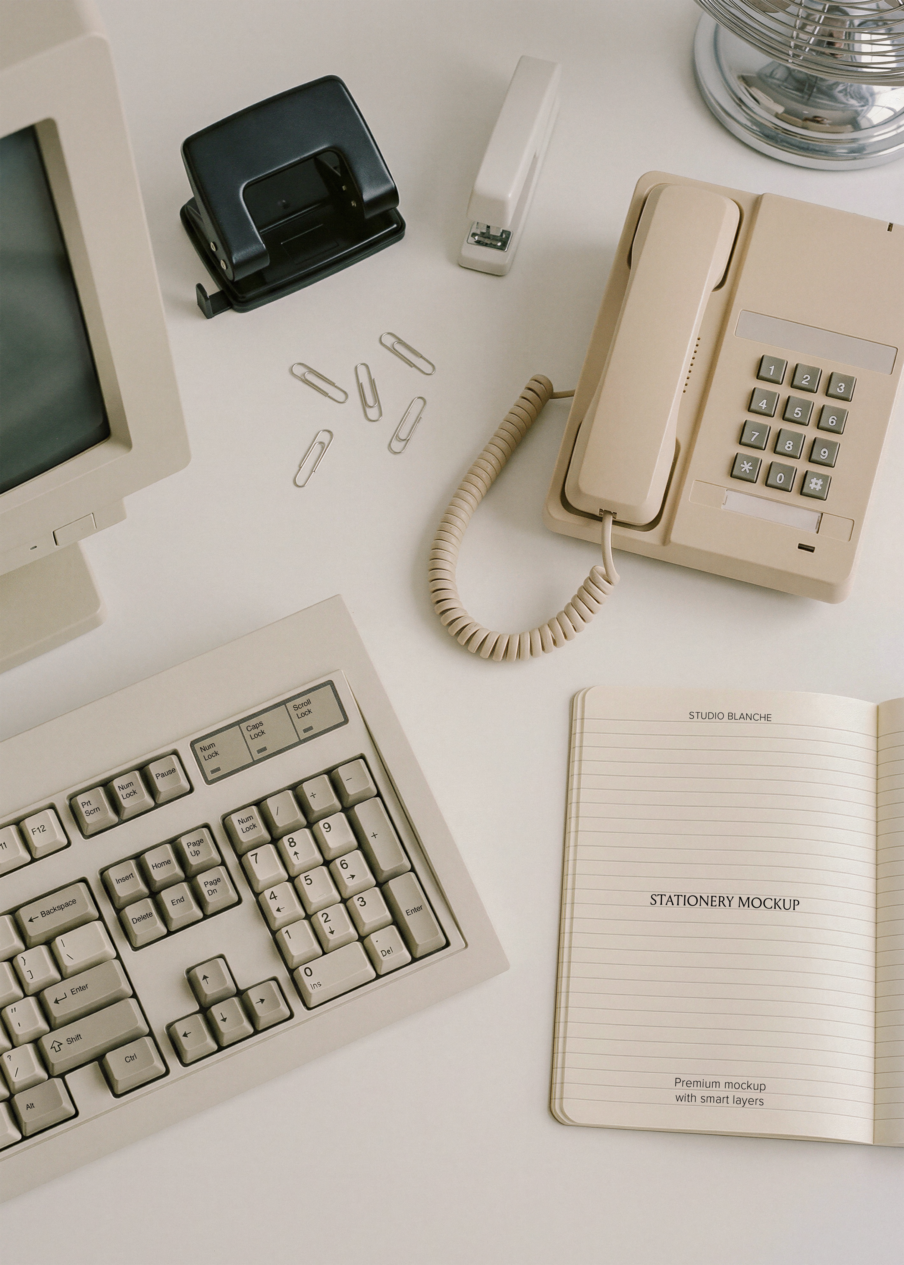 Ruled notebook spread on retro desk beside cream push-button phone, hole punch, stapler and scattered silver paper clips