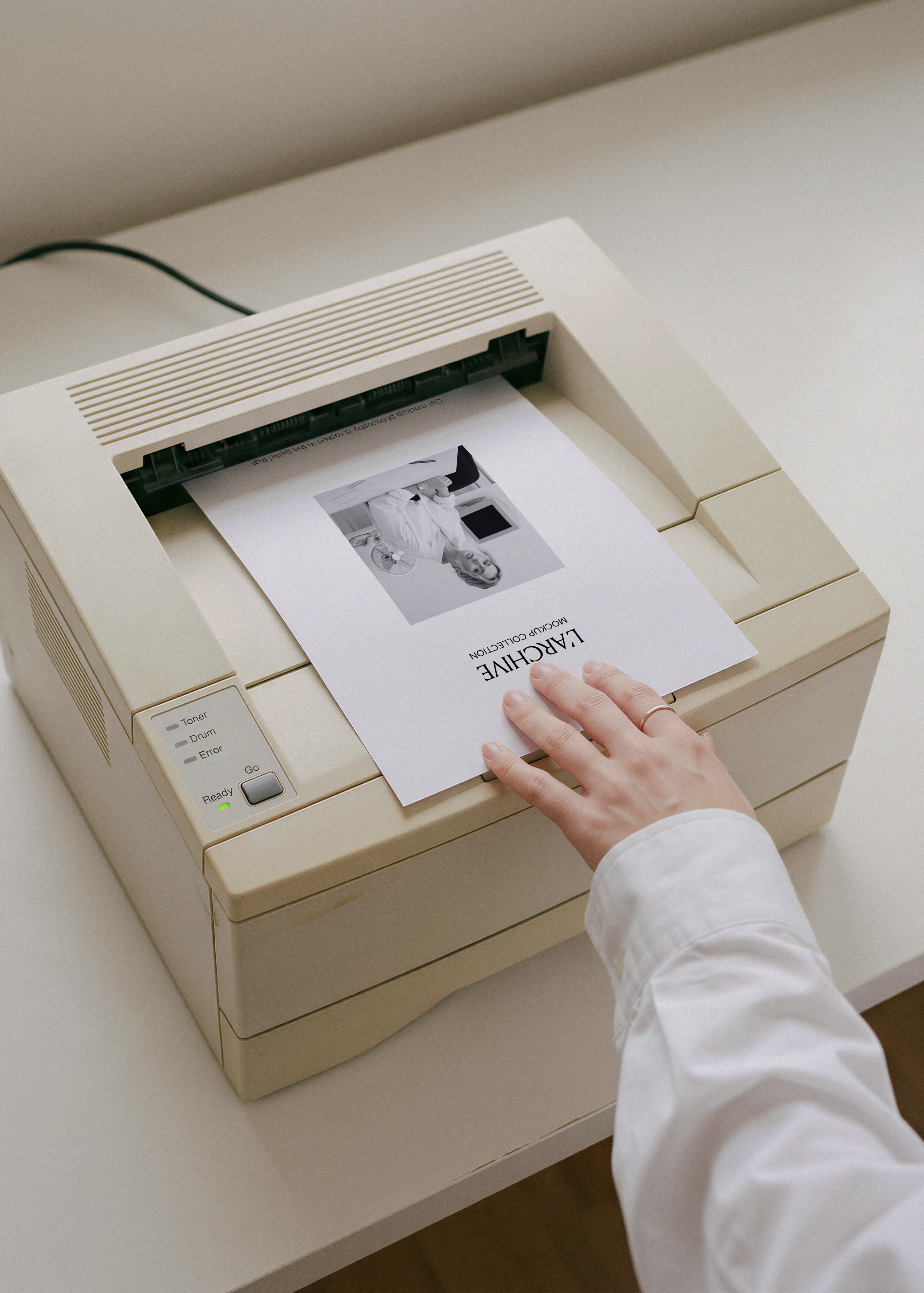 A4 card stationery mockup emerging from cream vintage laser printer, hand in white shirt guiding sheet, minimal desk scene