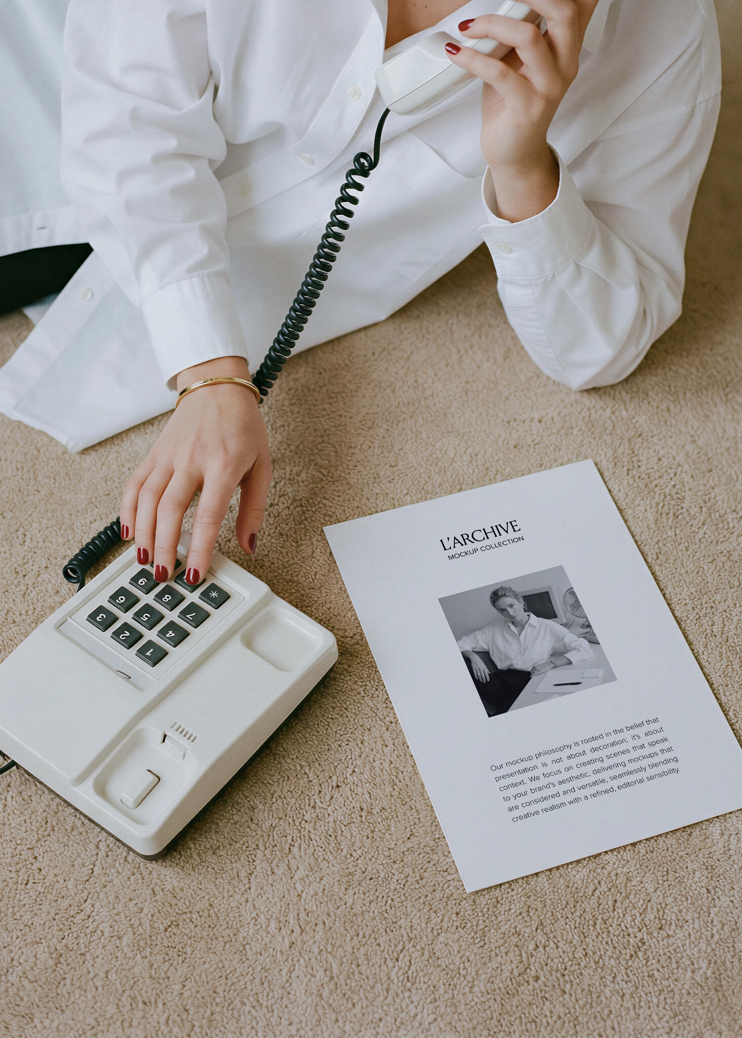 A4 card stationery mockup on beige carpet beside white retro push-button phone, hand dialling with red nails, gold bangle