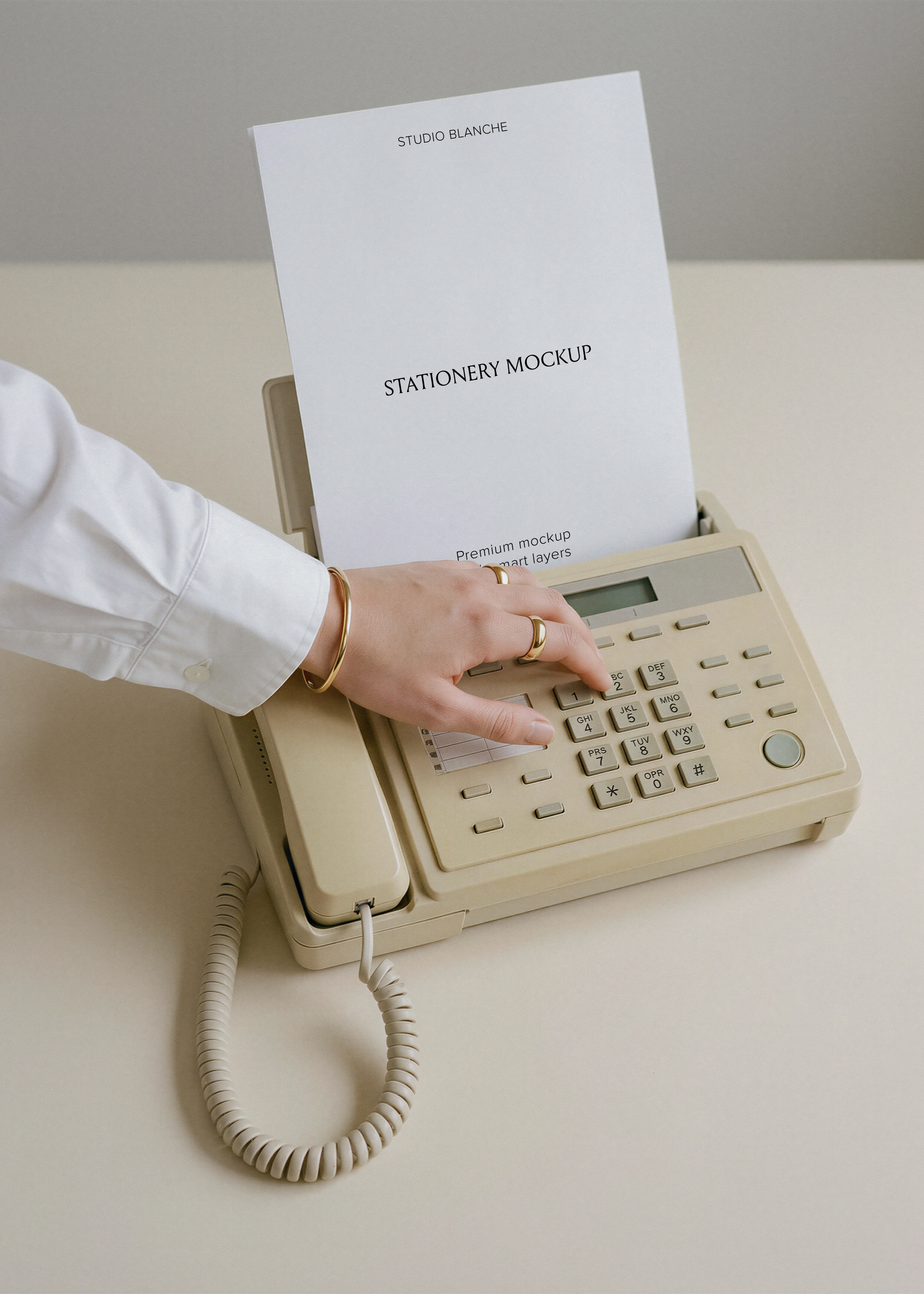 Blank A4 letterhead emerging from retro beige fax phone, coiled cord on pale desk, white shirt sleeve and gold jewellery
