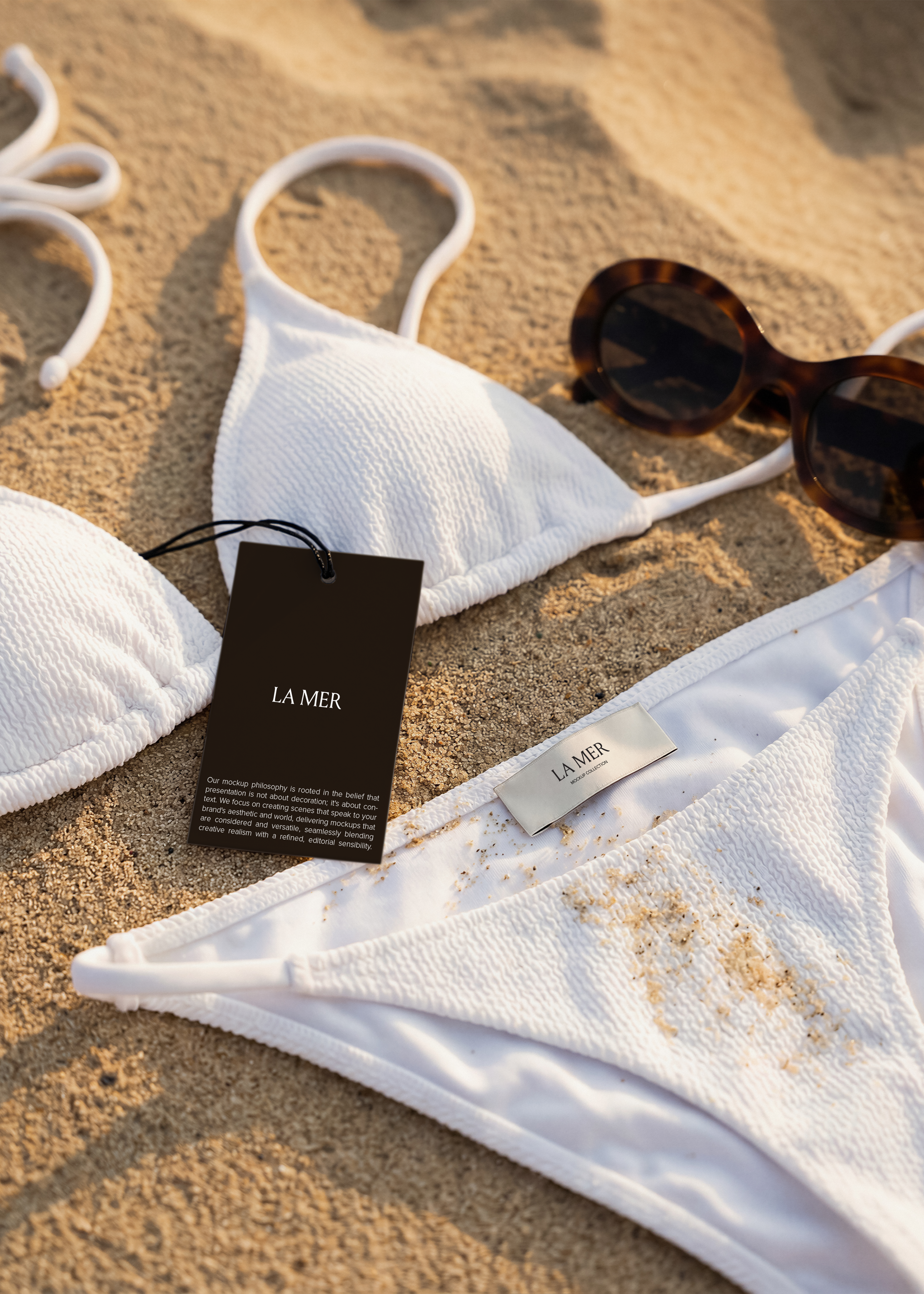 Dark swing tag mockup and silver garment label on white crinkle bikini laid flat on warm beach sand 