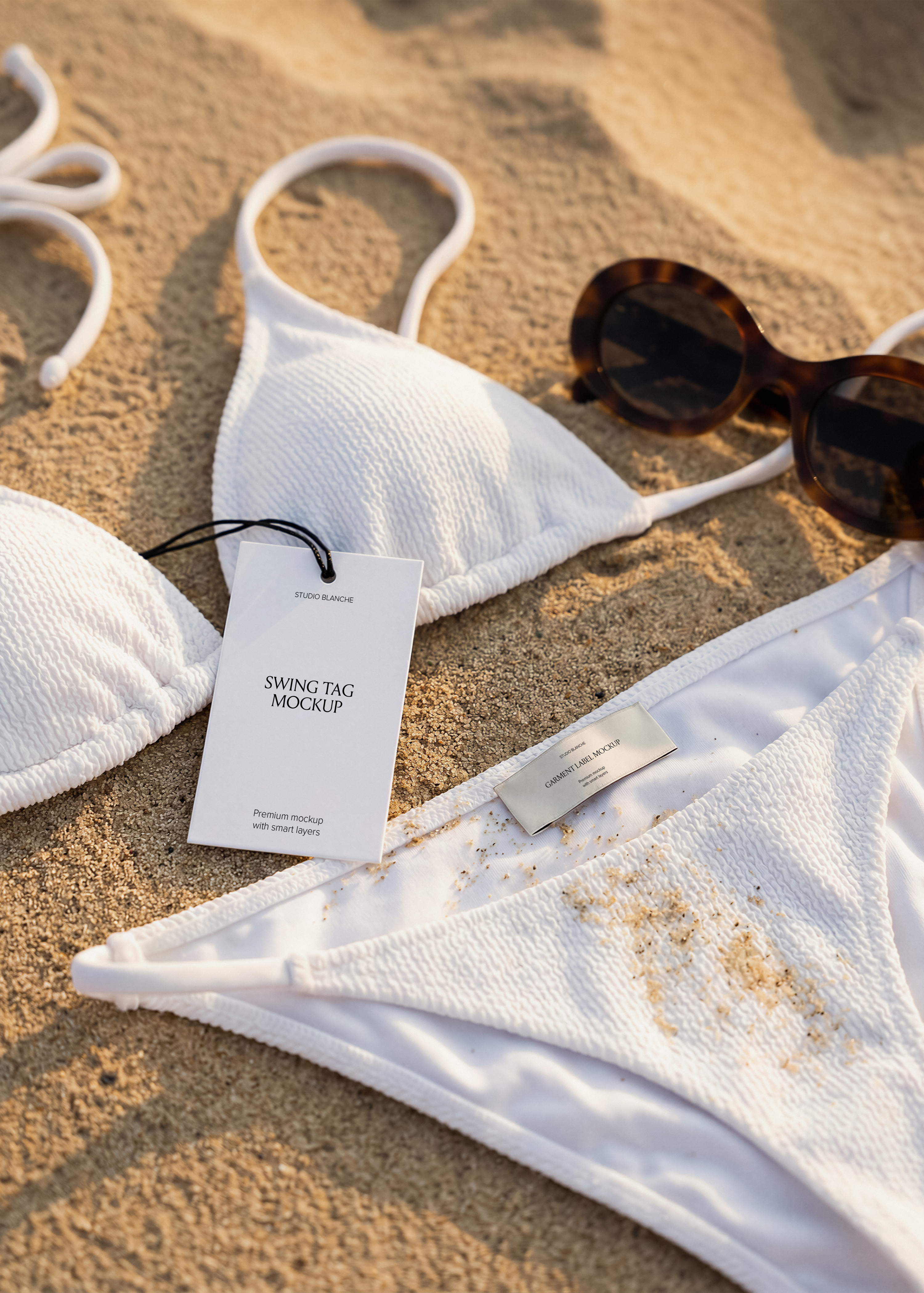 White swing tag and garment label on white crinkle bikini on beach sand, tortoiseshell sunglasses in scene