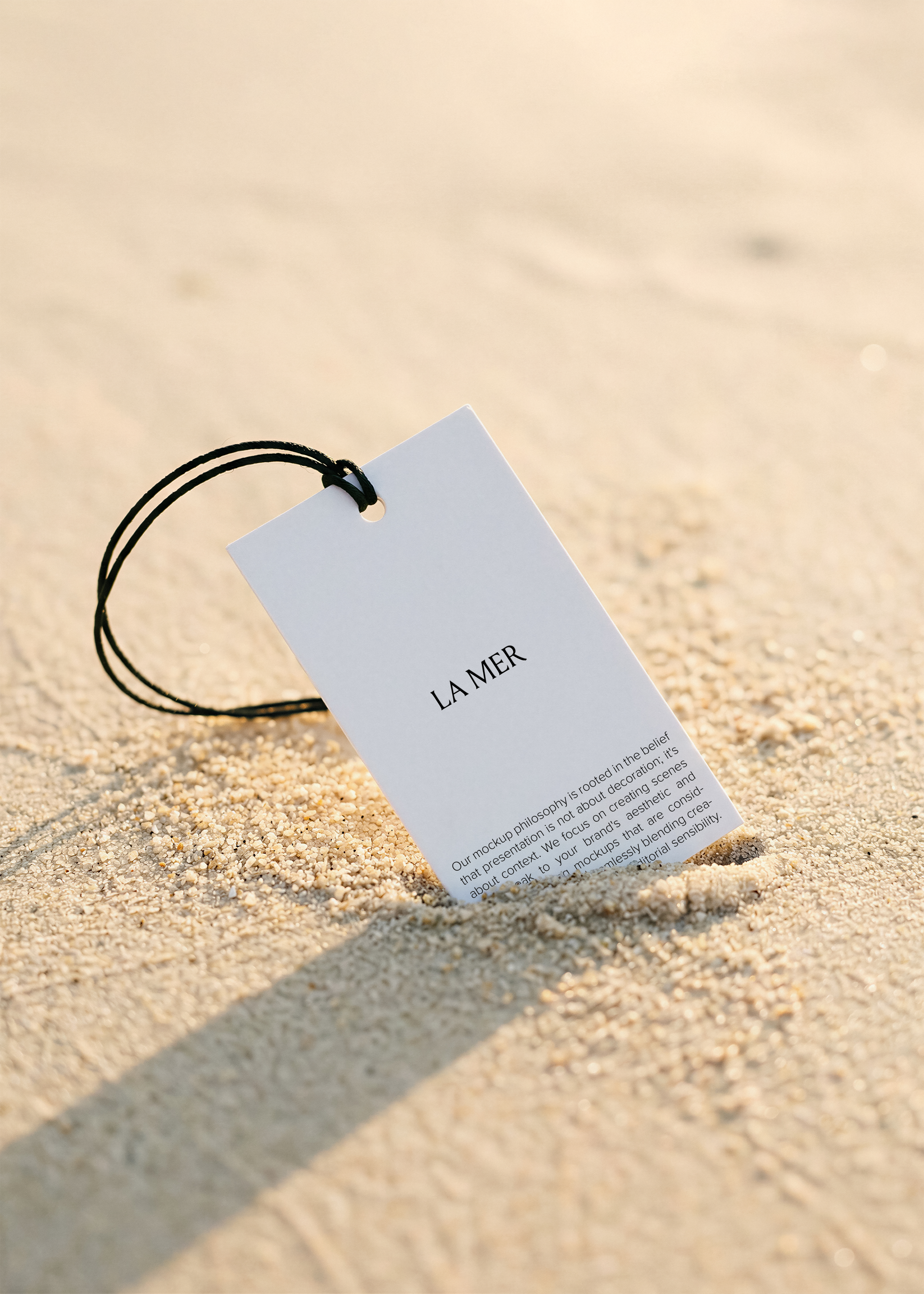 White swing tag with black cord standing upright in fine beach sand, warm golden light, soft shadow 