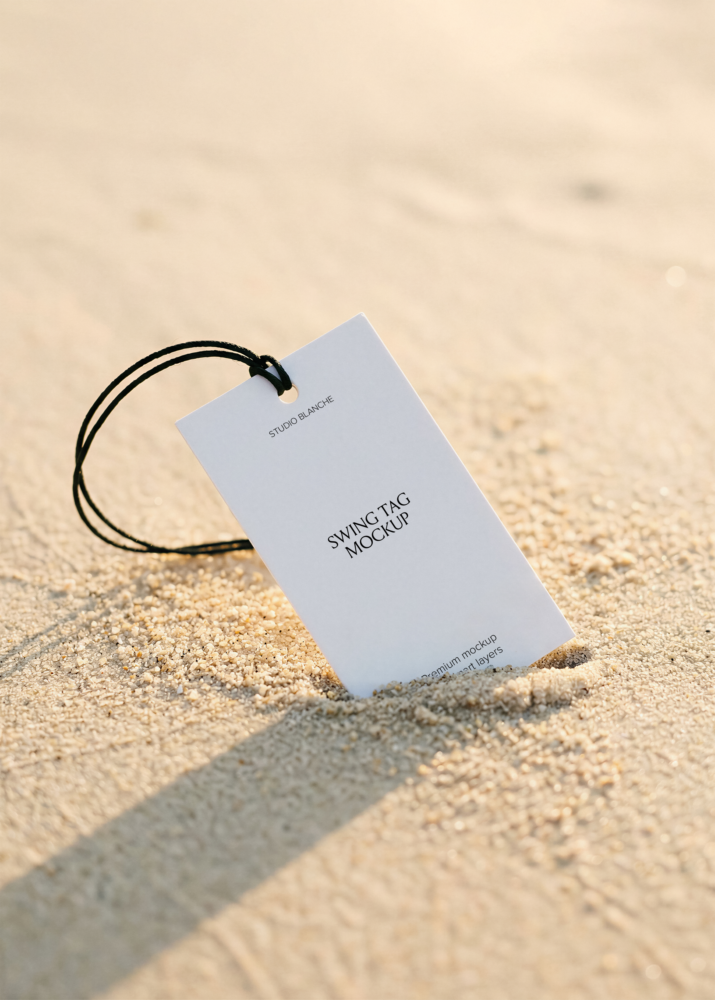White hang tag mockup propped in beach sand with black string, bleached sand background, editorial styling