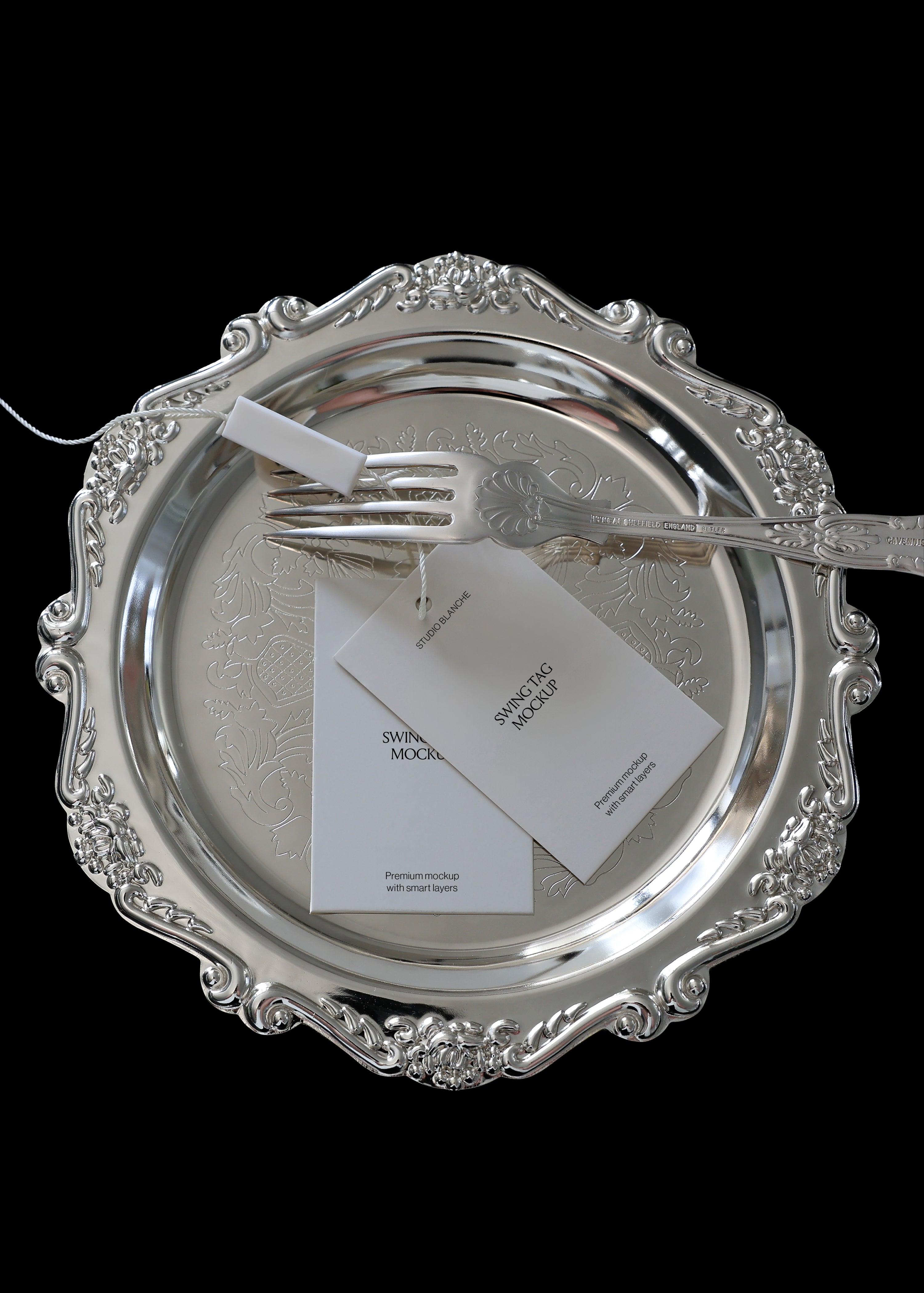 Swing tag mockup with white string resting on an ornate silver plate with engraved fork, black background