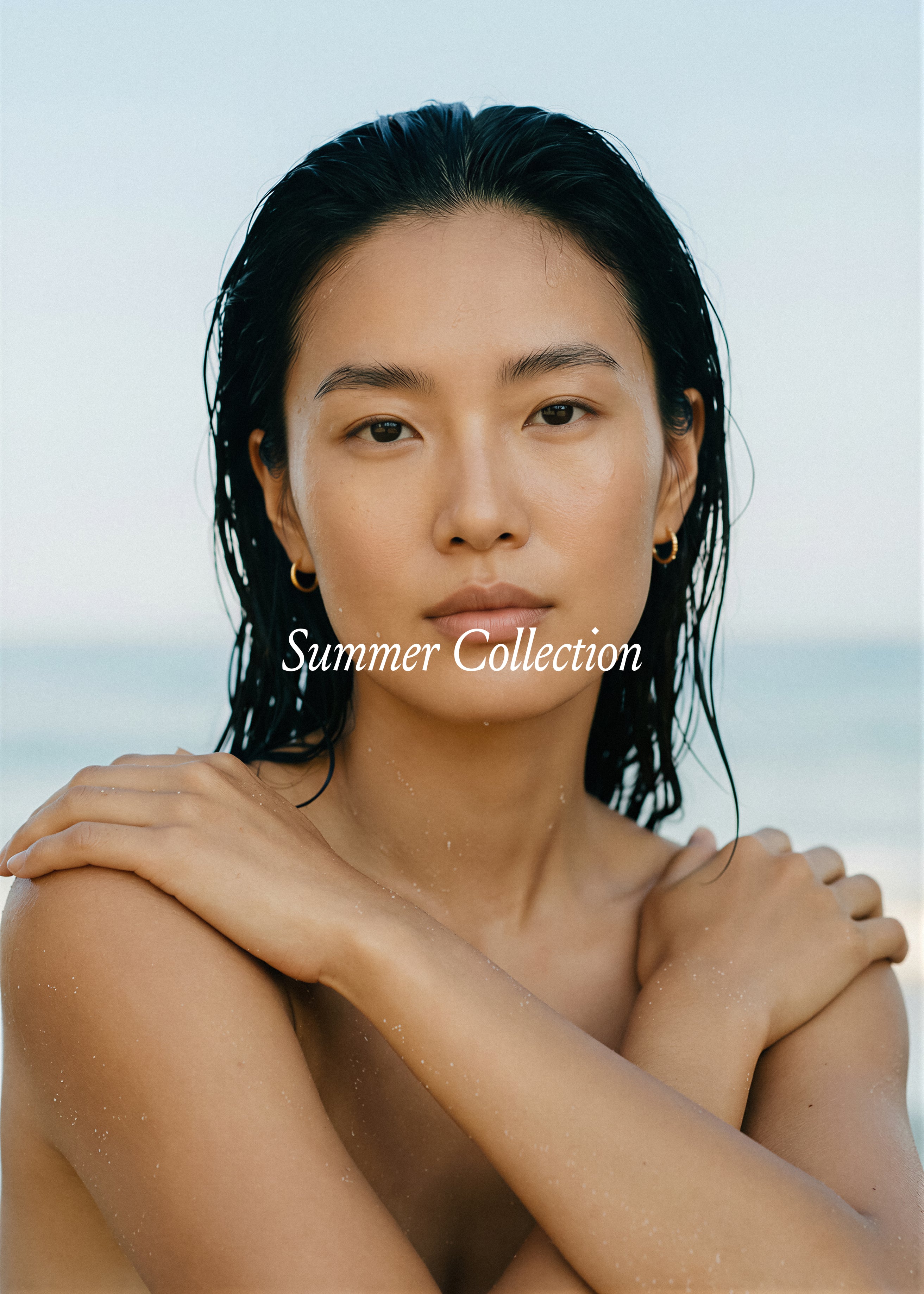 Dark-haired model, arms crossed, wet hair, gold hoops, cool ocean background. Summer Collection Vol. II, Studio Blanche.