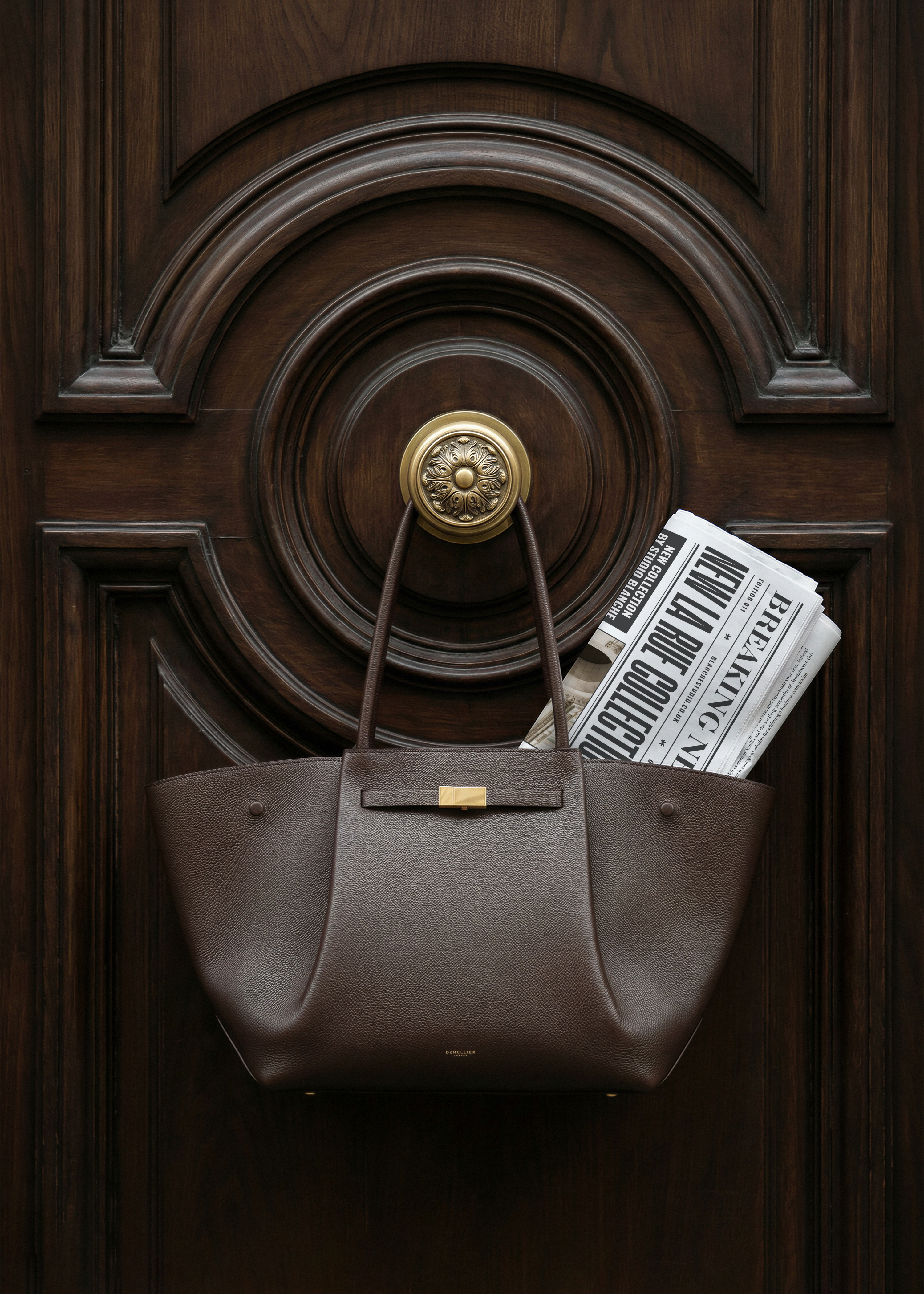 Newspaper mockup with editorial headline in dark leather Demellier tote bag on ornate wooden door, from Studio Blanche