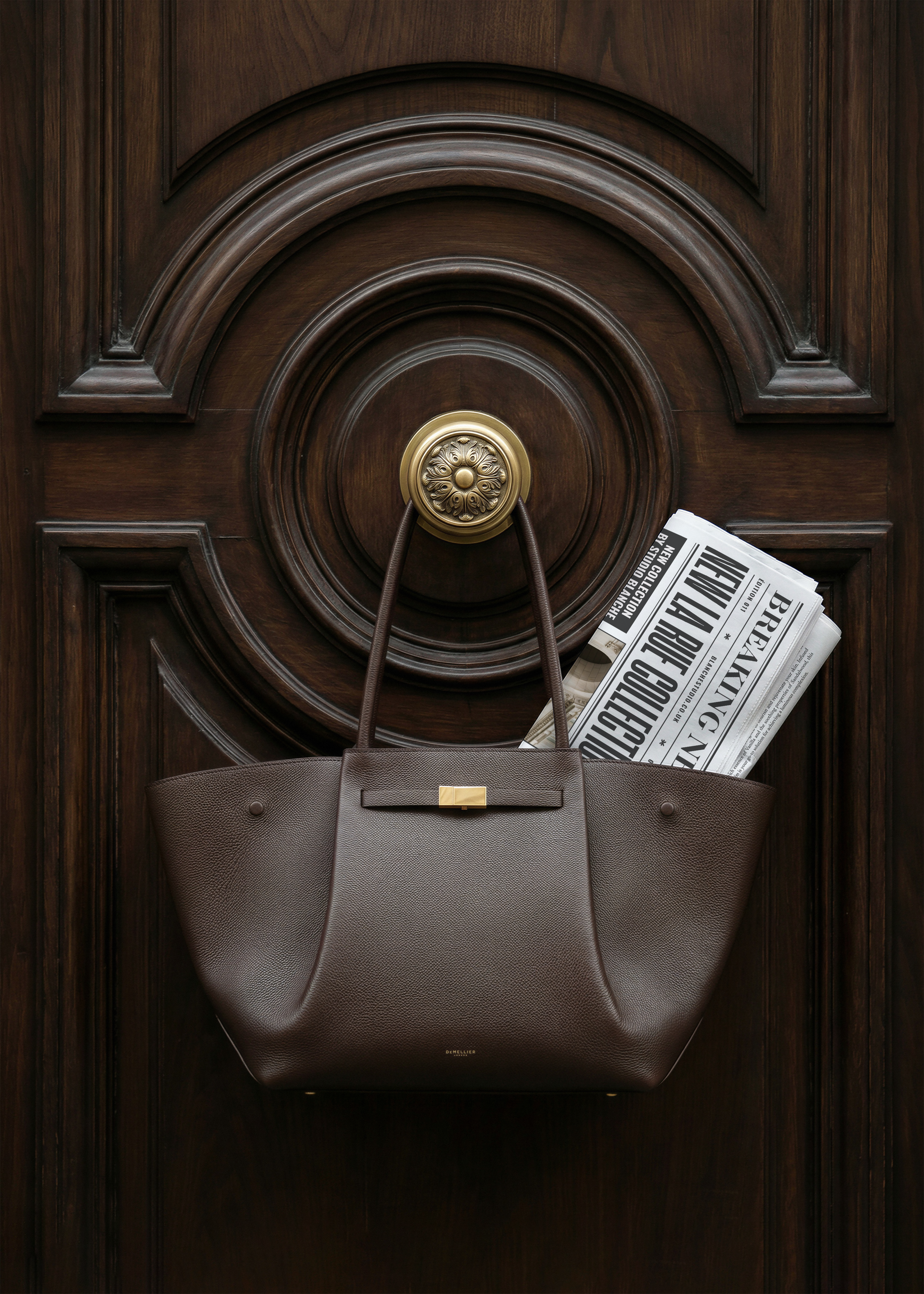 Newspaper mockup with editorial headline in dark leather Demellier tote bag on ornate wooden door, from Studio Blanche