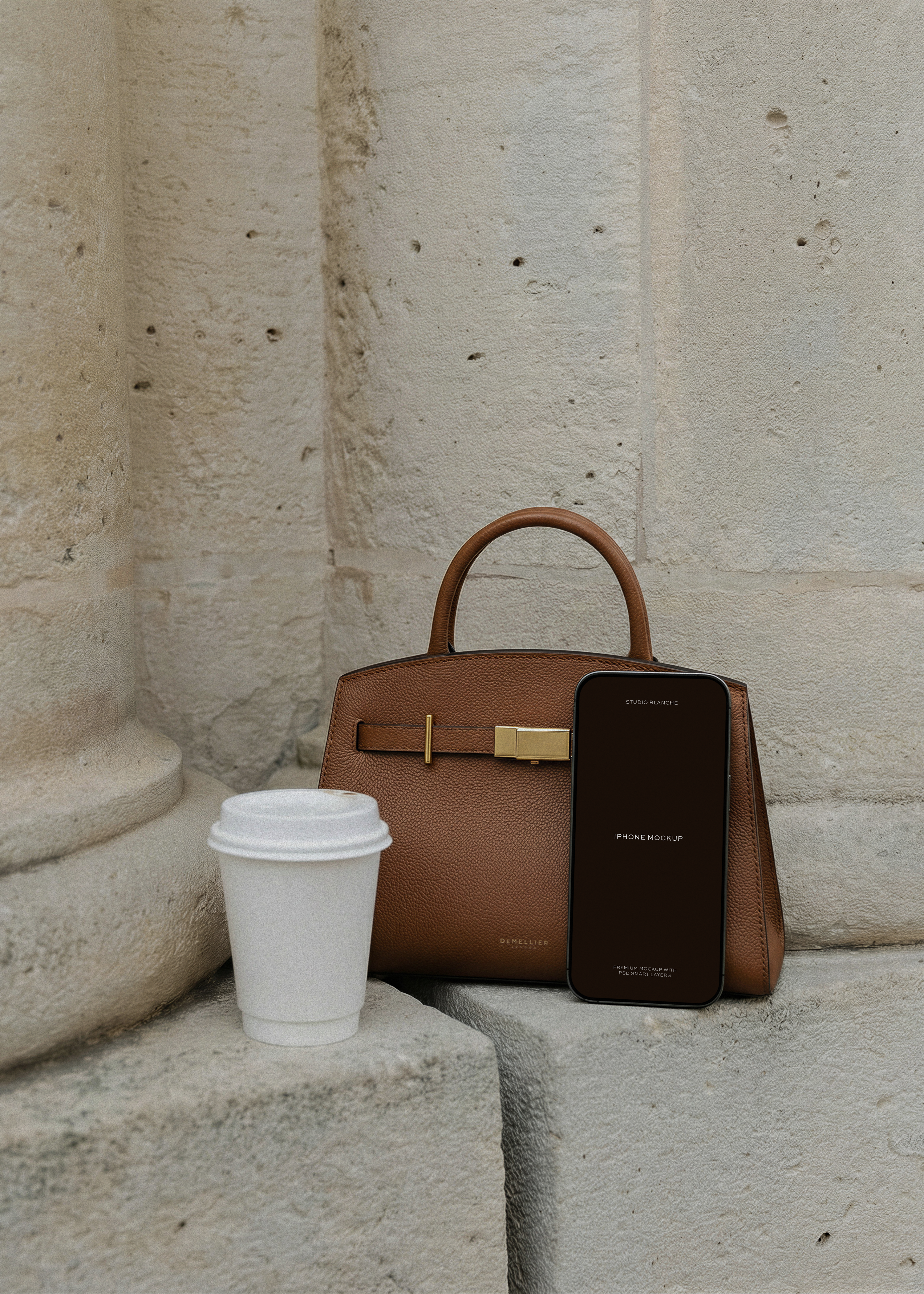 iPhone mockup against tan leather handbag beside a stone column with coffee cup, from Studio Blanche