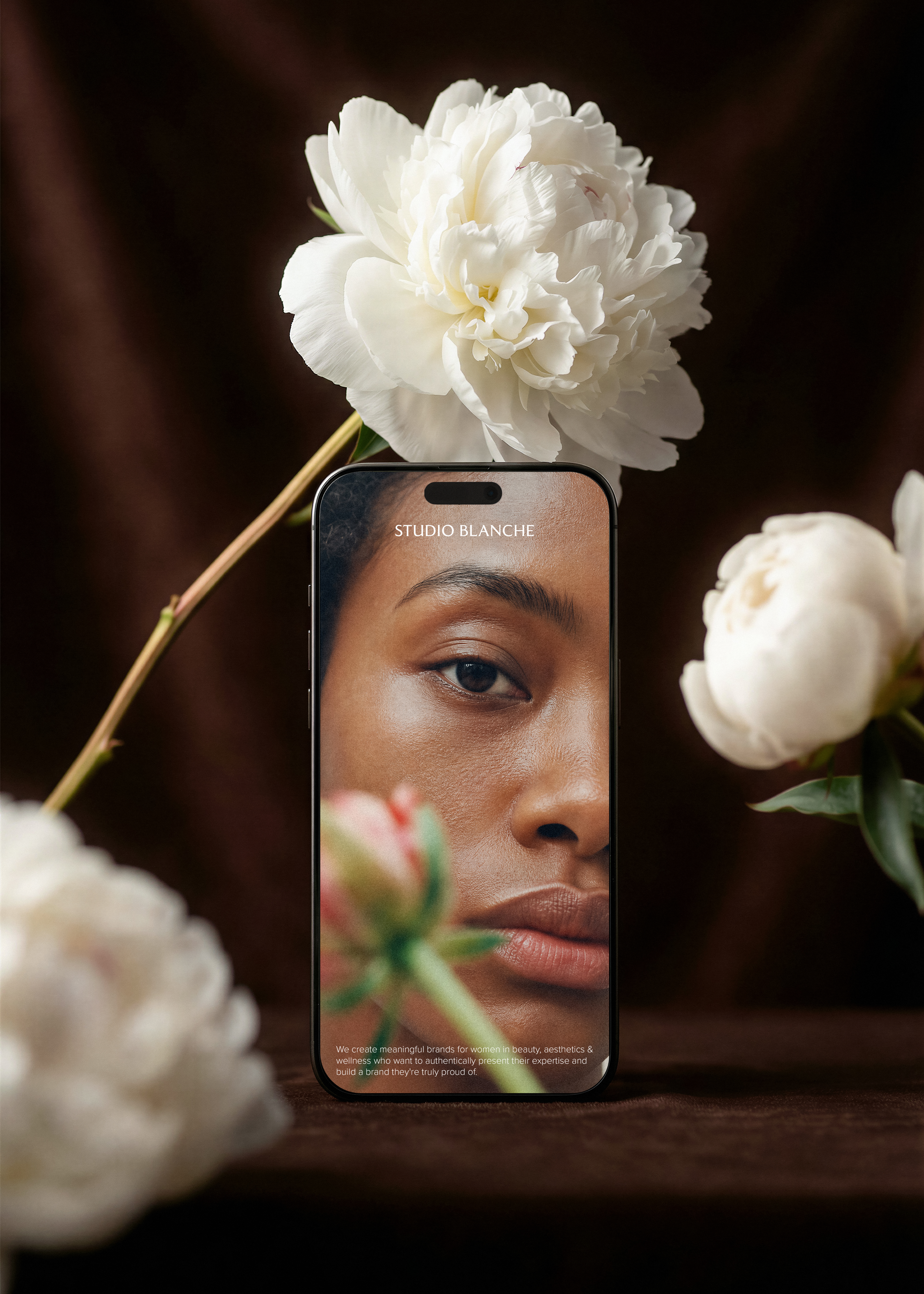 iPhone mockup displaying a beauty brand website surrounded by white peonies on a dark brown background, Studio Blanche