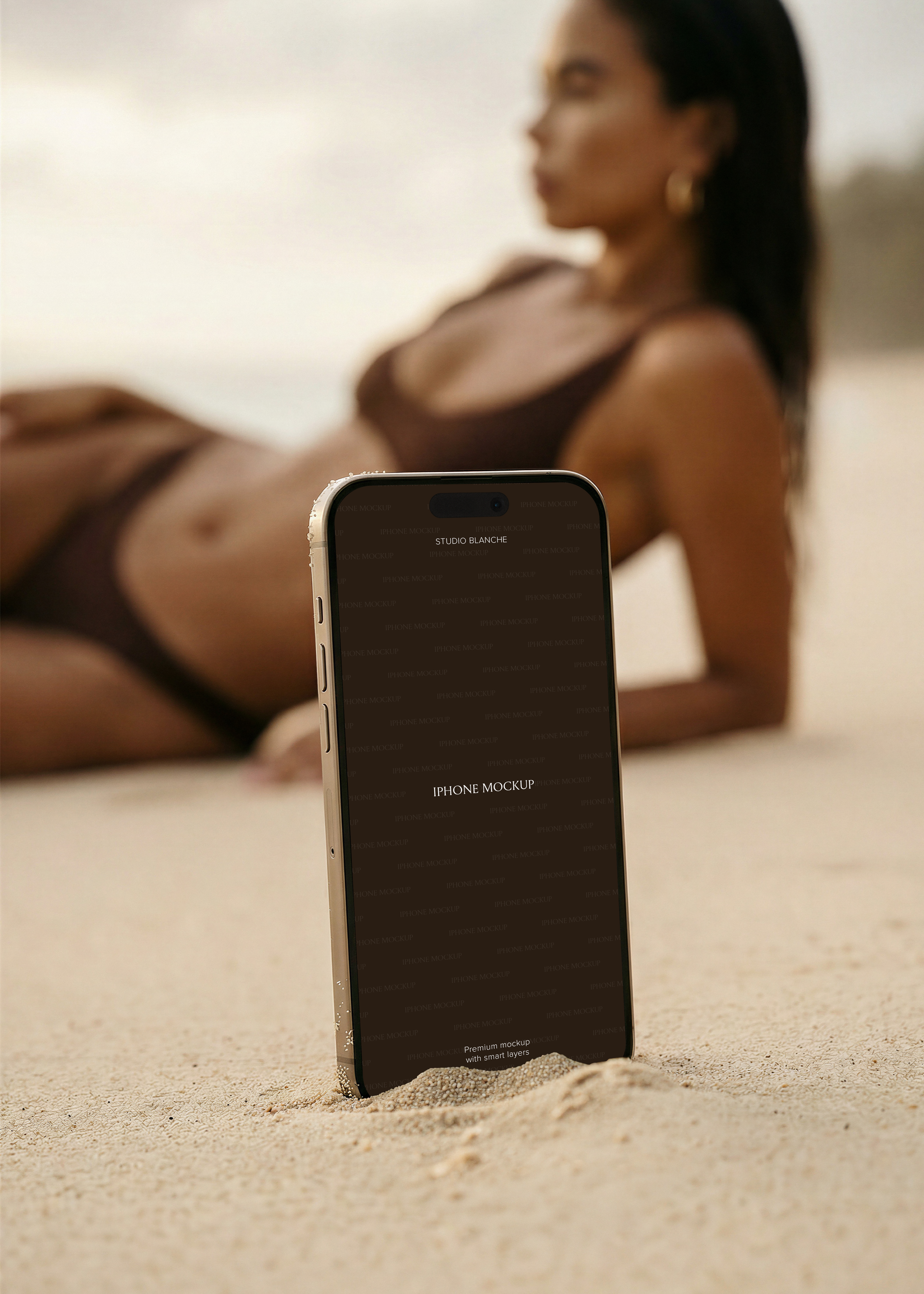 Gold iPhone propped upright in pale dry sand, woman in dark swimwear reclining in soft focus behind, warm tones