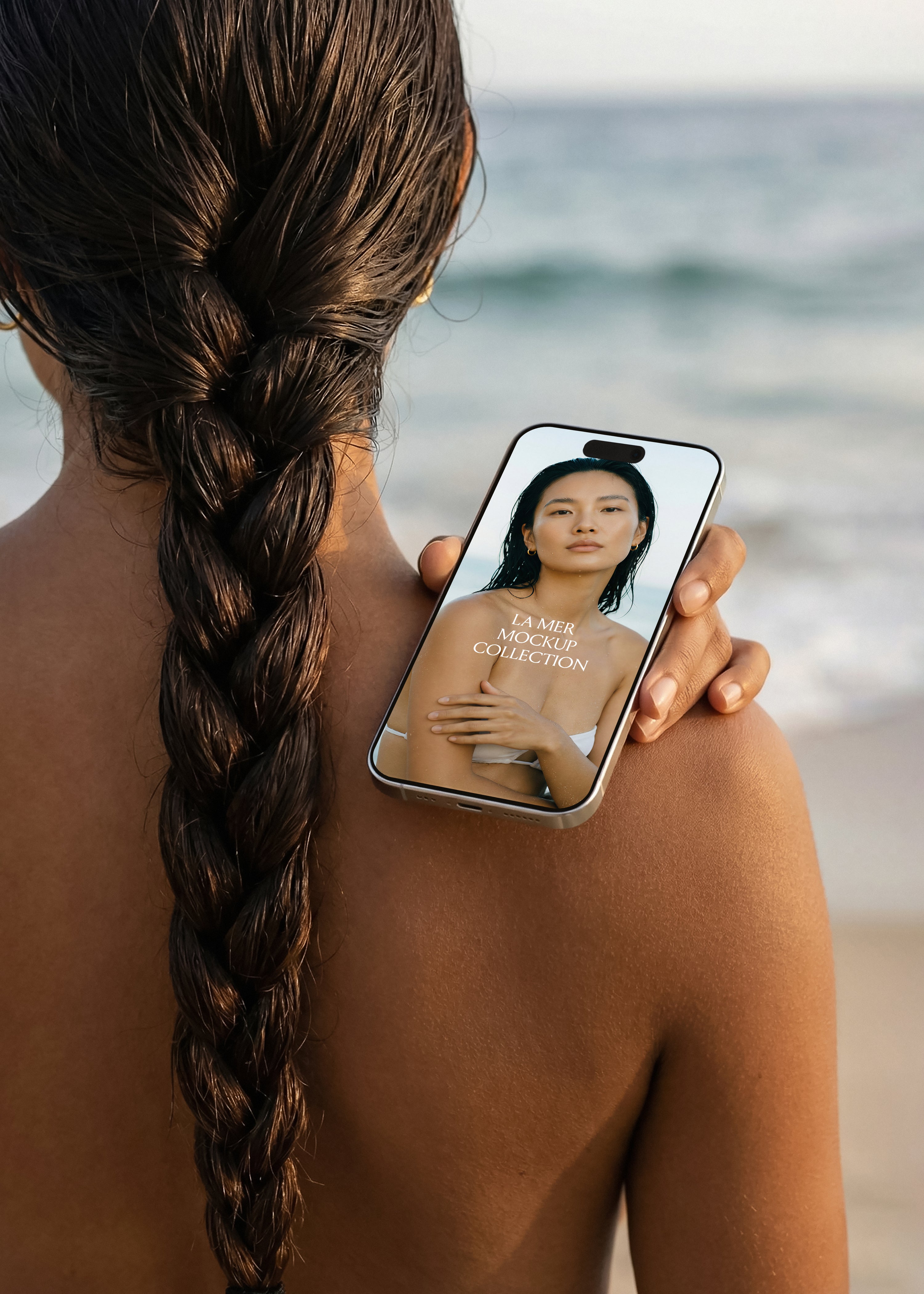 Model with braided wet hair facing ocean holding iPhone mockup over shoulder, soft coastal light, La Mer collection