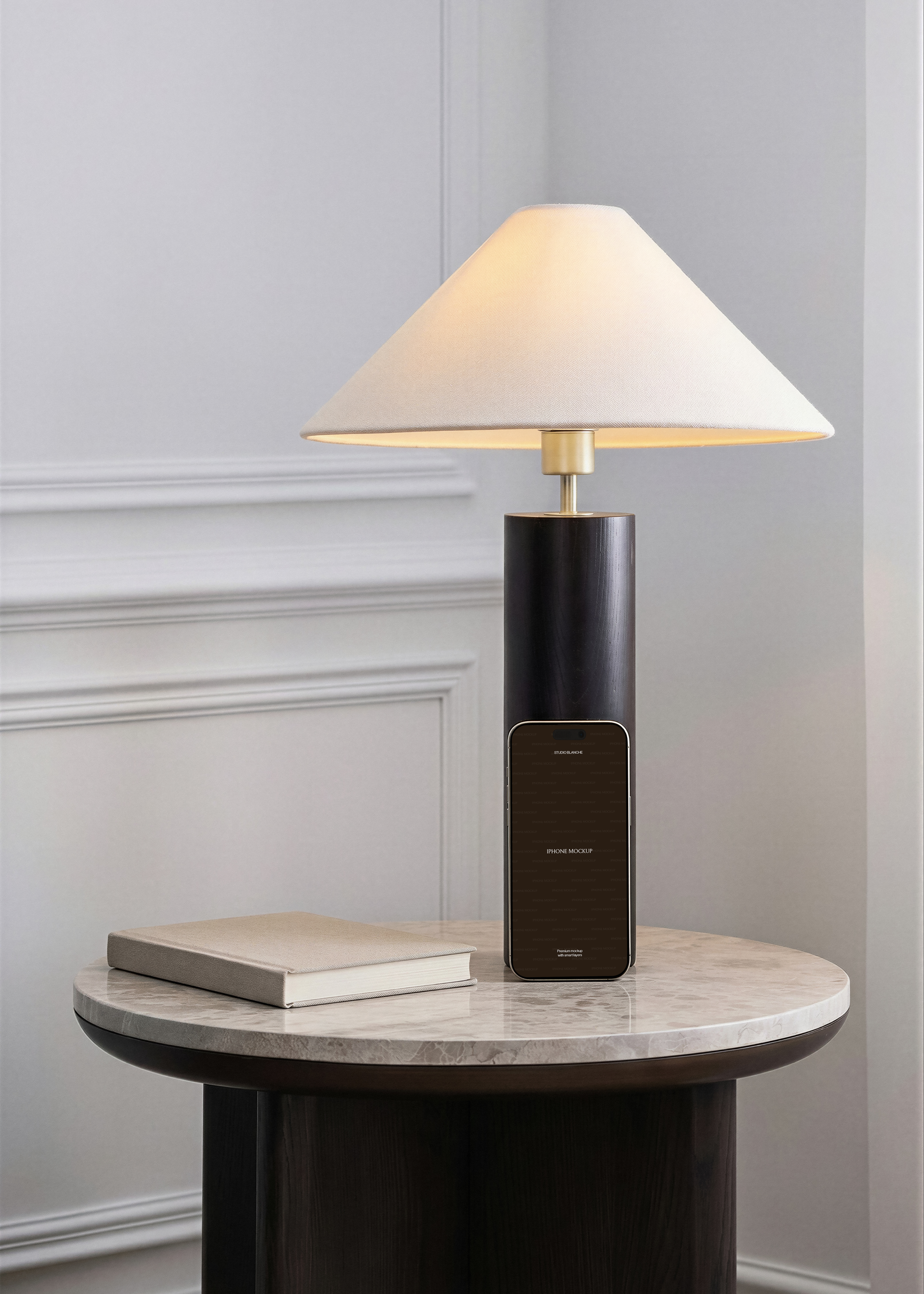 iPhone mockup leaning against dark cylindrical brass lamp on marble-top side table, dove-grey panelled wall interior