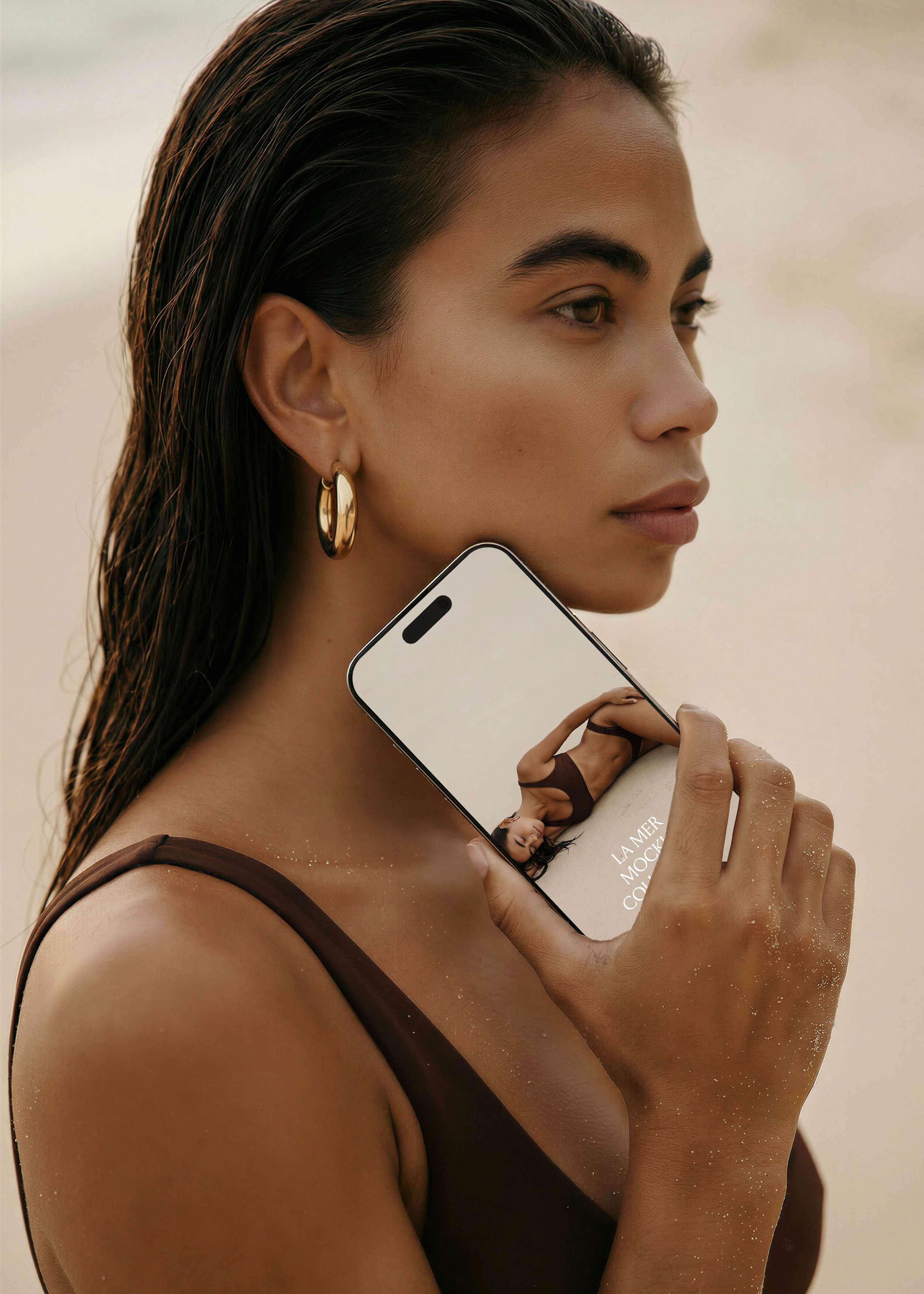 Model in profile holding iPhone mockup to jaw, wet hair, gold hoop earring, sandy beach background, La Mer collection