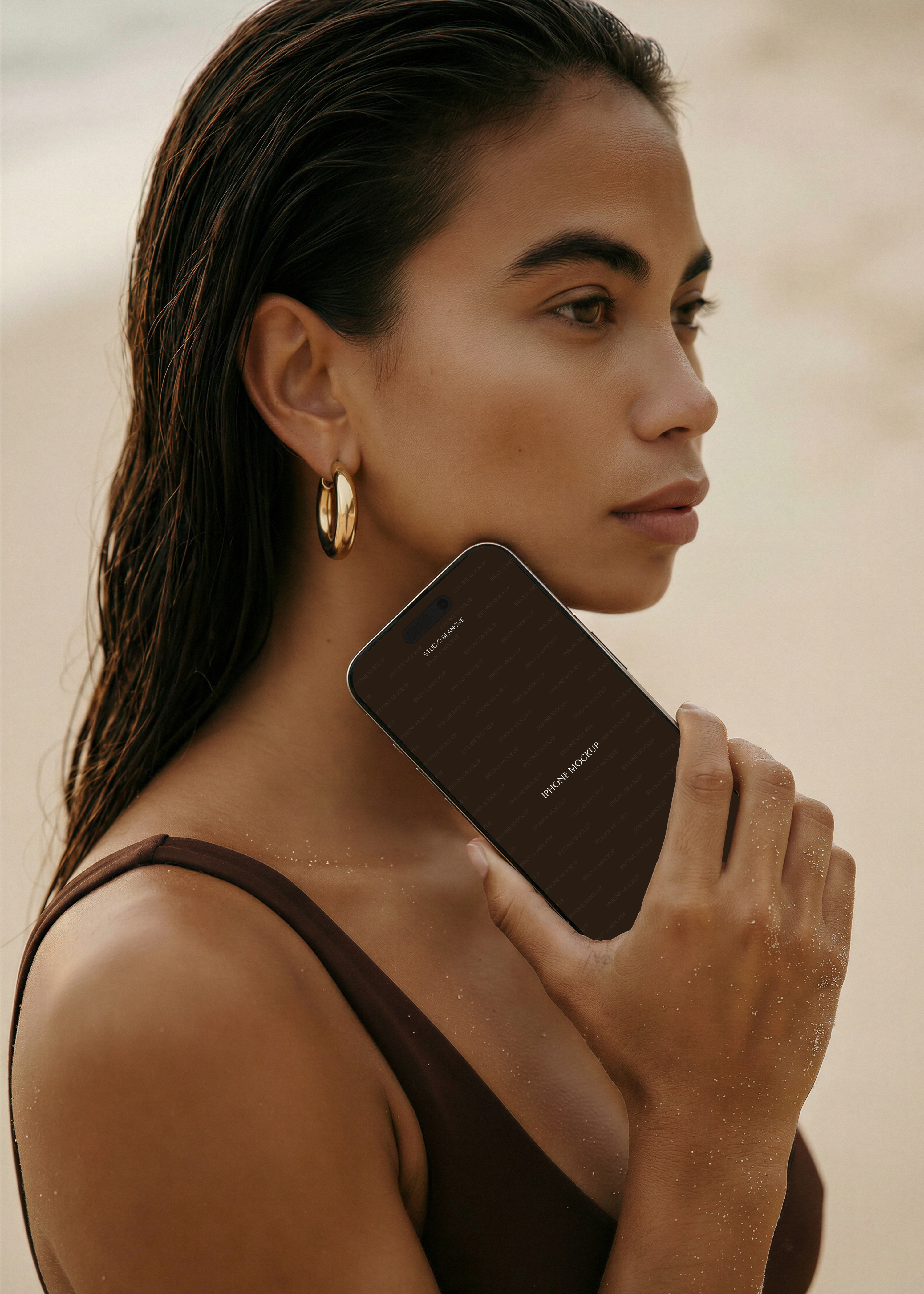 Beach iPhone mockup held against model's jaw in profile, dark swimwear, sand-dusted skin, warm editorial light