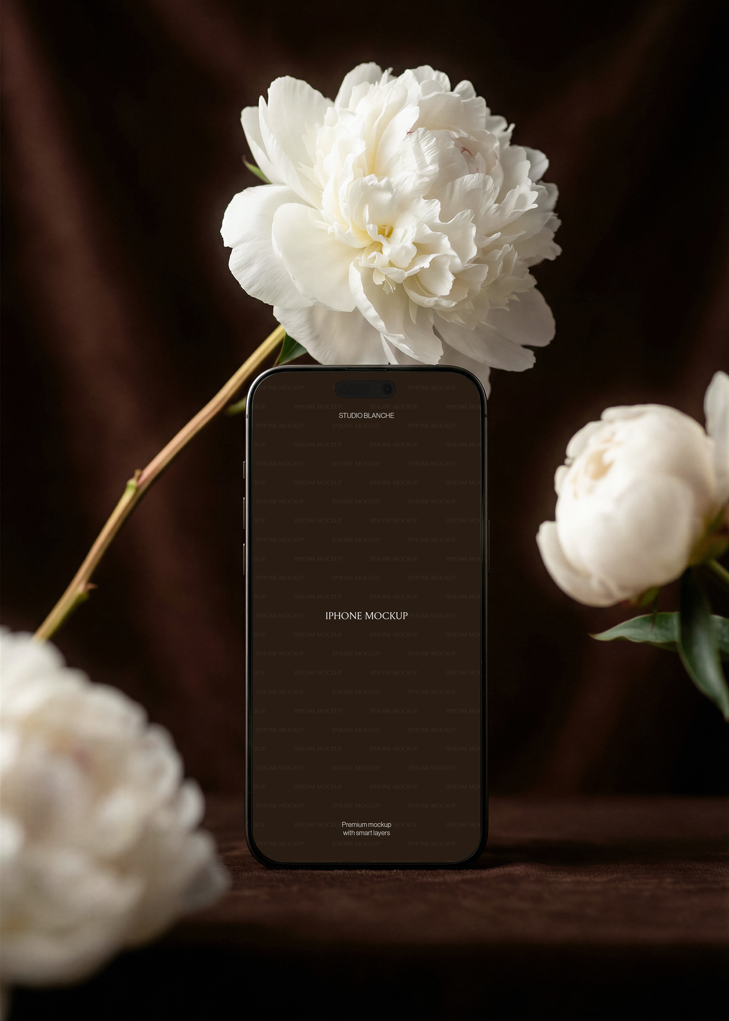 iPhone mockup showing screen area, large white peony overhead on stem, blurred peony blooms left and right, dark brown velvet surface 