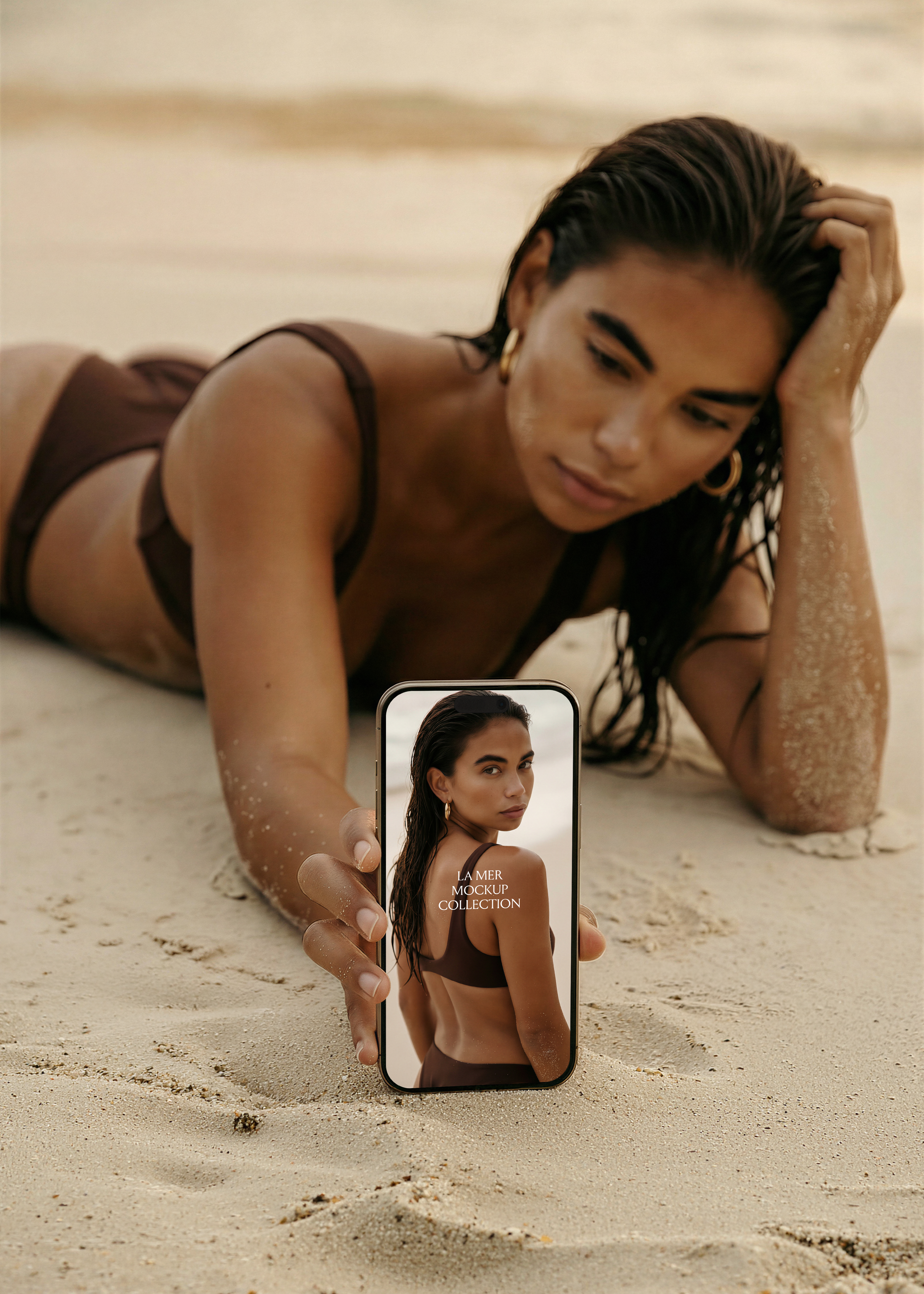 Woman in dark bikini lying on sandy beach holding iPhone mockup facing camera, warm editorial lighting, La Mer collection