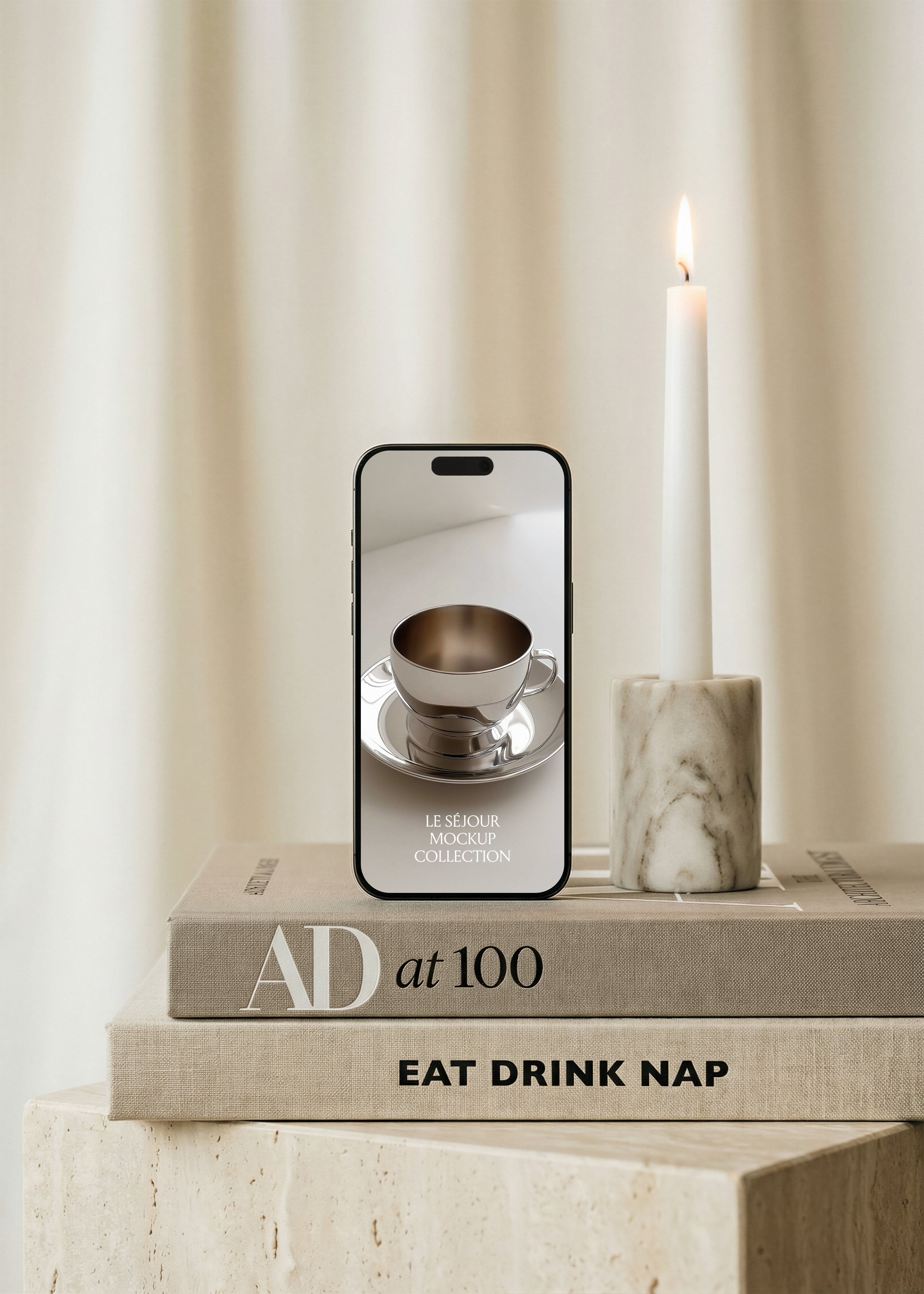 iPhone mockup standing upright on stacked coffee table books on a travertine plinth beside a lit candle in marble holder